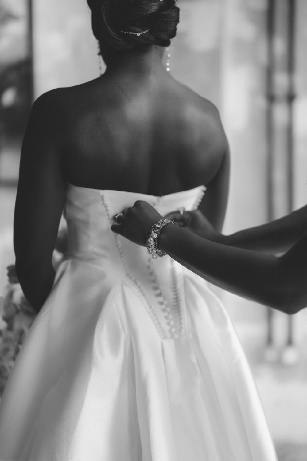 Black and white photo of a bride in a strapless gown being buttoned up by a bridesmaid wearing a crystal bracelet.