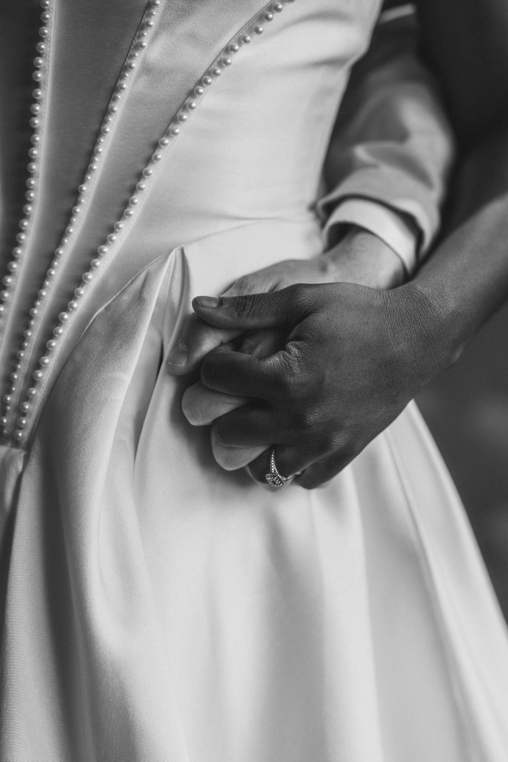 Black and white photo of interracial couple holding hands, bride wearing wedding dress with pearl details and engagement ring.
