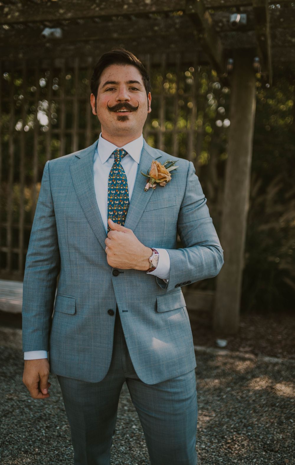 Groom in light blue suit with floral boutonniere and patterned tie, posing outdoors with stylish mustache.