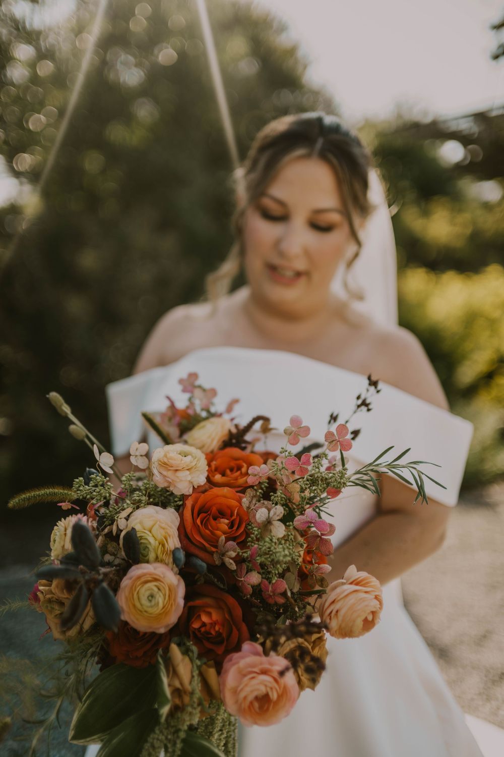 Bride in white strapless gown holding vibrant bouquet of orange, pink, and peach ranunculus with greenery outdoors.