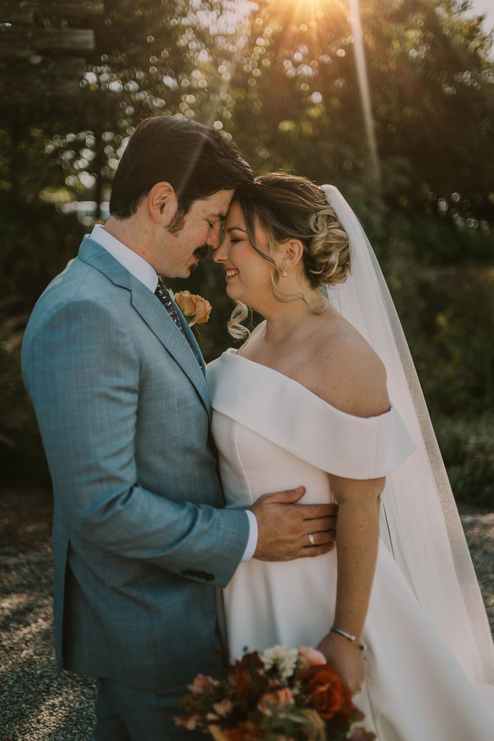 A bride and groom share a romantic moment at golden hour, with the bride in a white off-shoulder gown and groom in a blue suit.