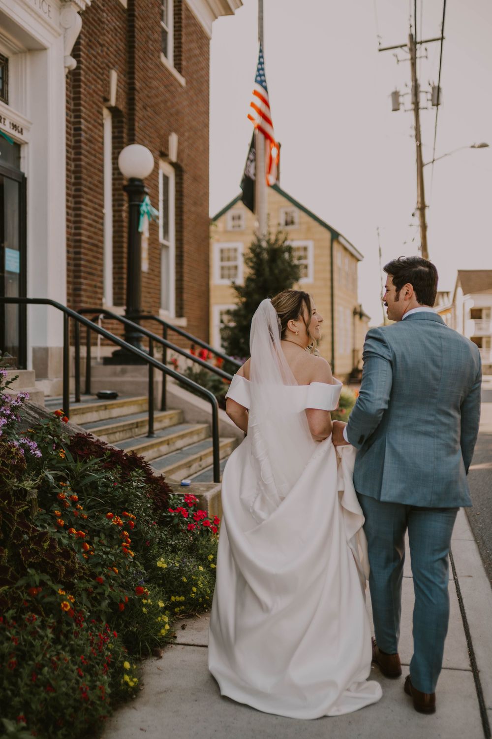 A bride and groom walk hand in hand along a sidewalk past a brick building with an American flag on their wedding day.