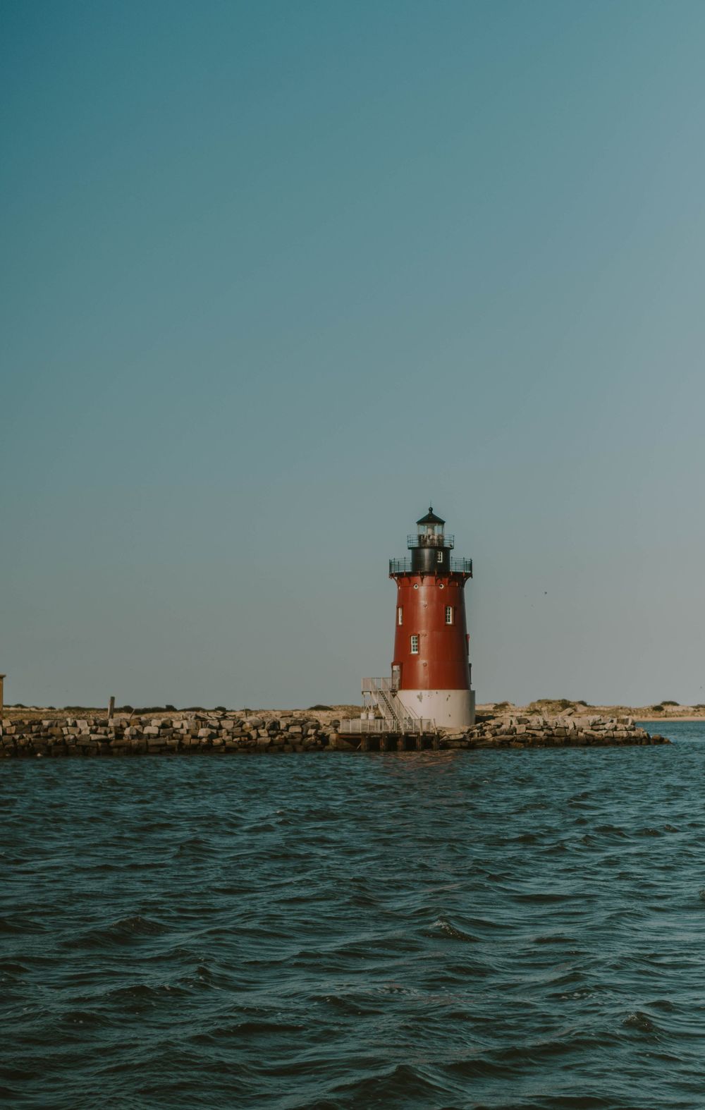 Red and white lighthouse standing on a rocky breakwater surrounded by calm ocean waters under a clear blue sky.