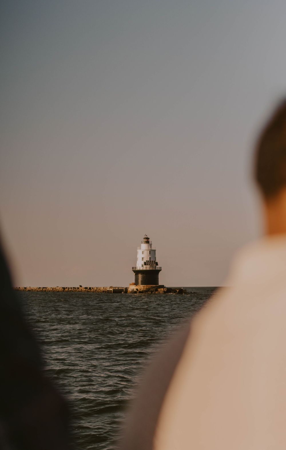 Two people view a historic white lighthouse standing on a rocky breakwater surrounded by calm ocean waters at golden hour.