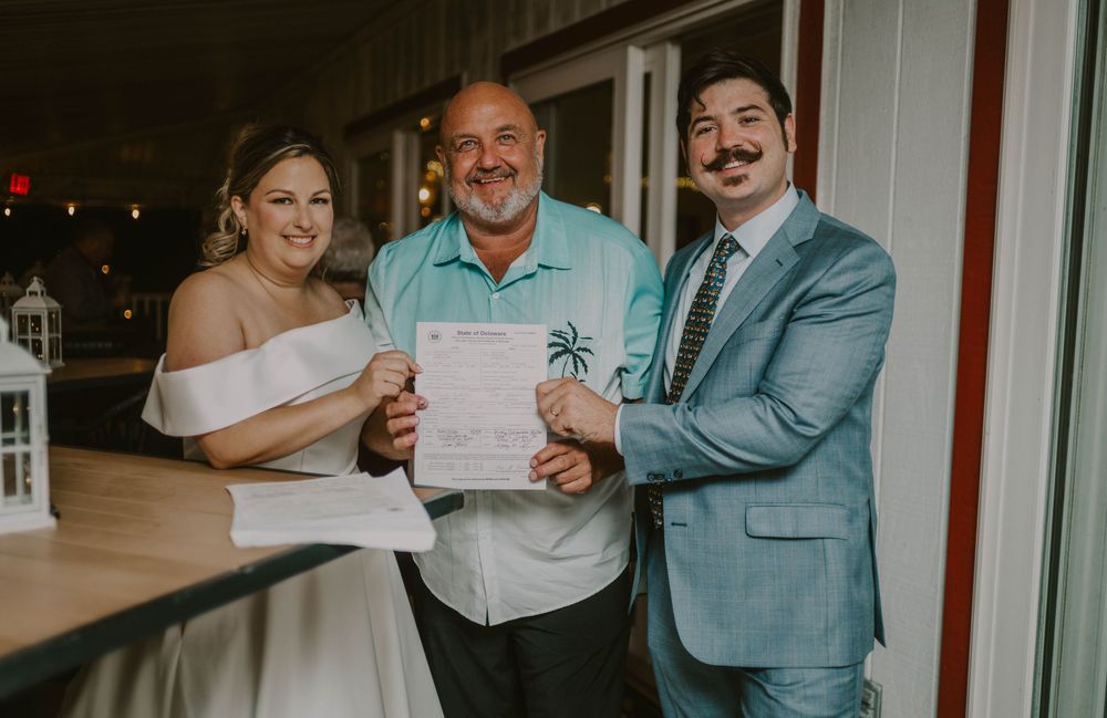 Bride and groom holding marriage certificate with officiant at outdoor wedding ceremony venue.