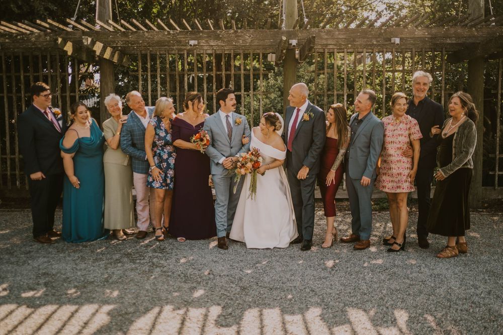 Wedding party poses together outdoors under a pergola, bride and groom centered among family members in formal attire.