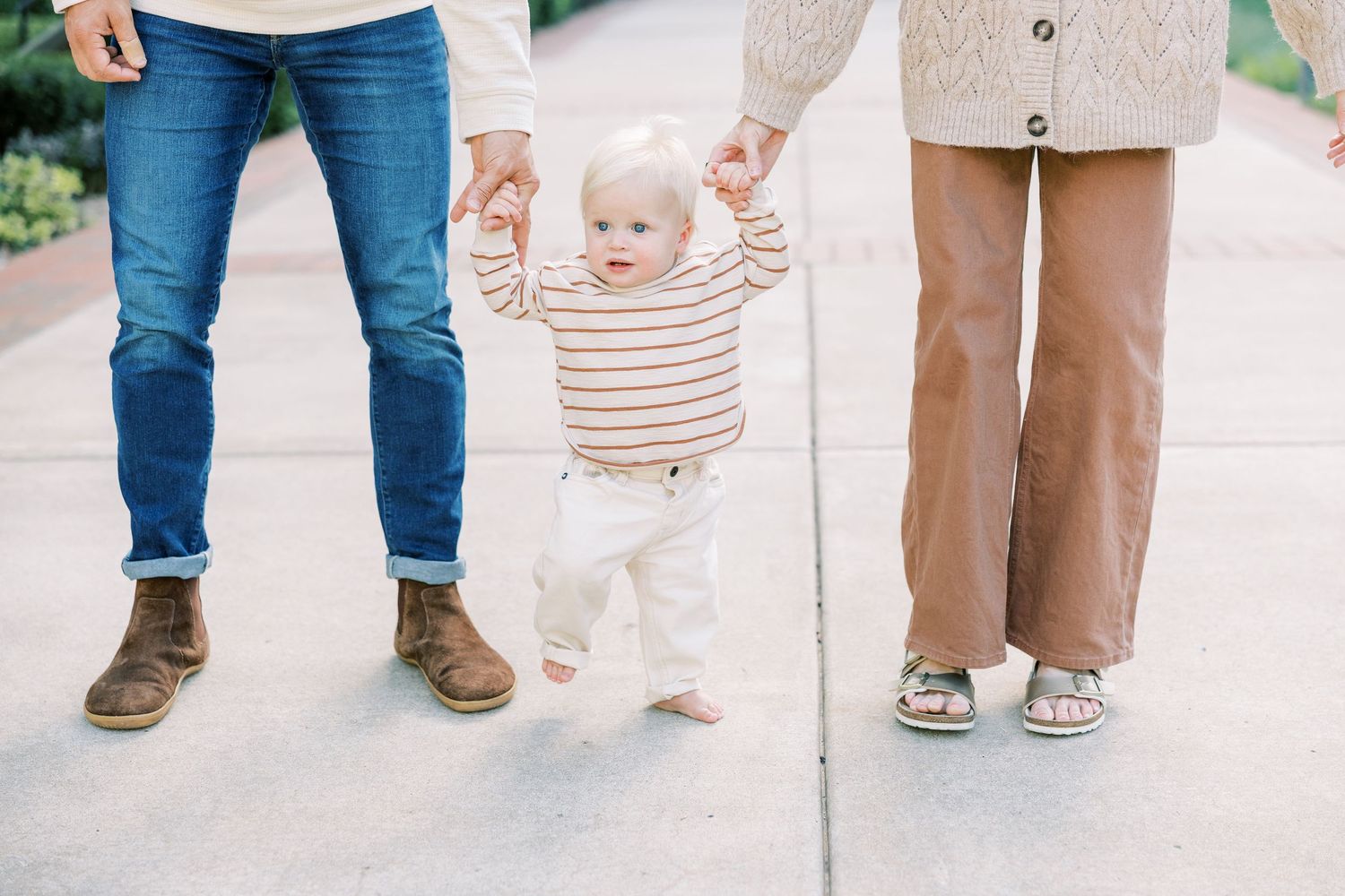 Baby taking first steps while parents hold hands outdoors on a sunny day.