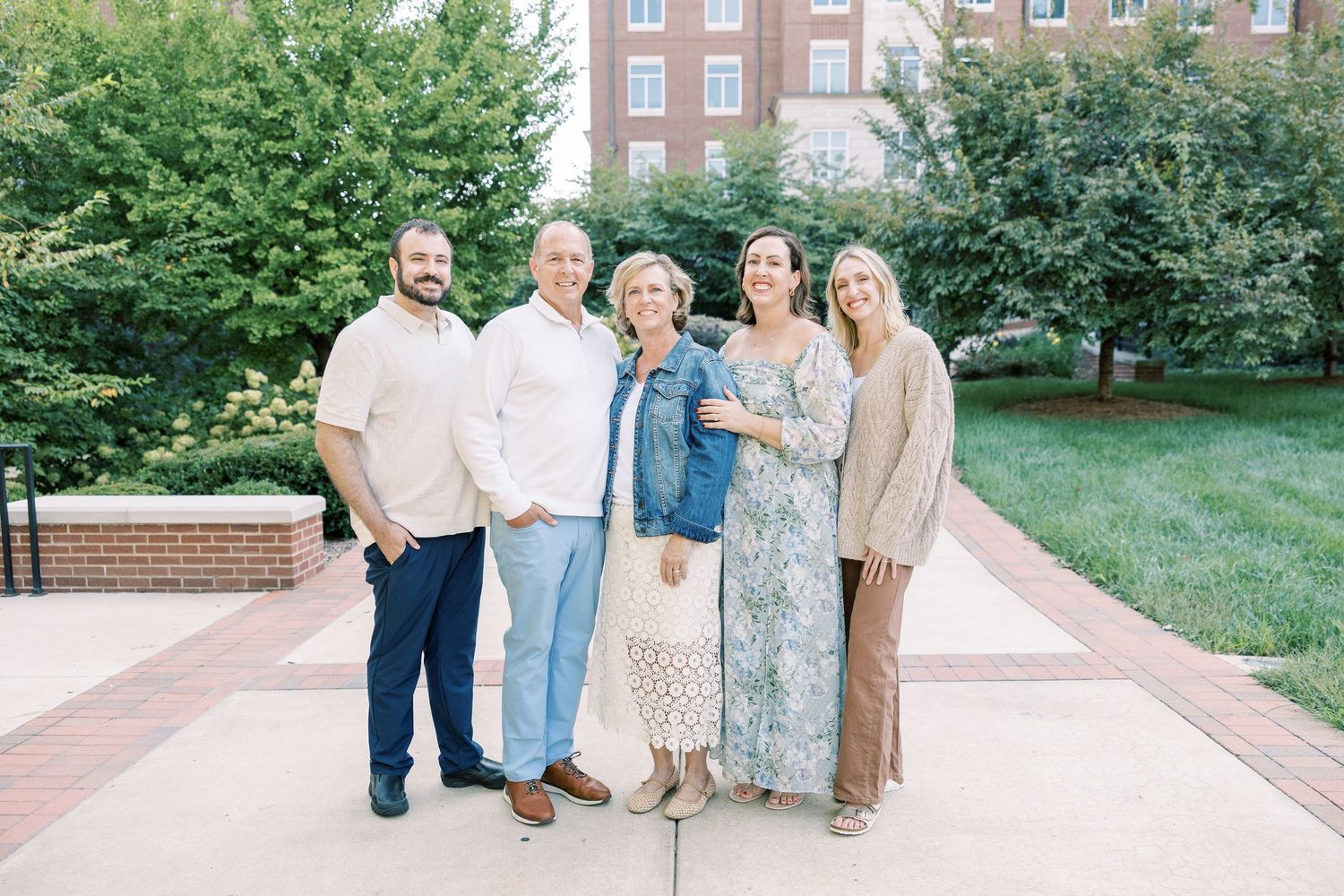 Family of five adults smiling together outdoors on a brick pathway near greenery.