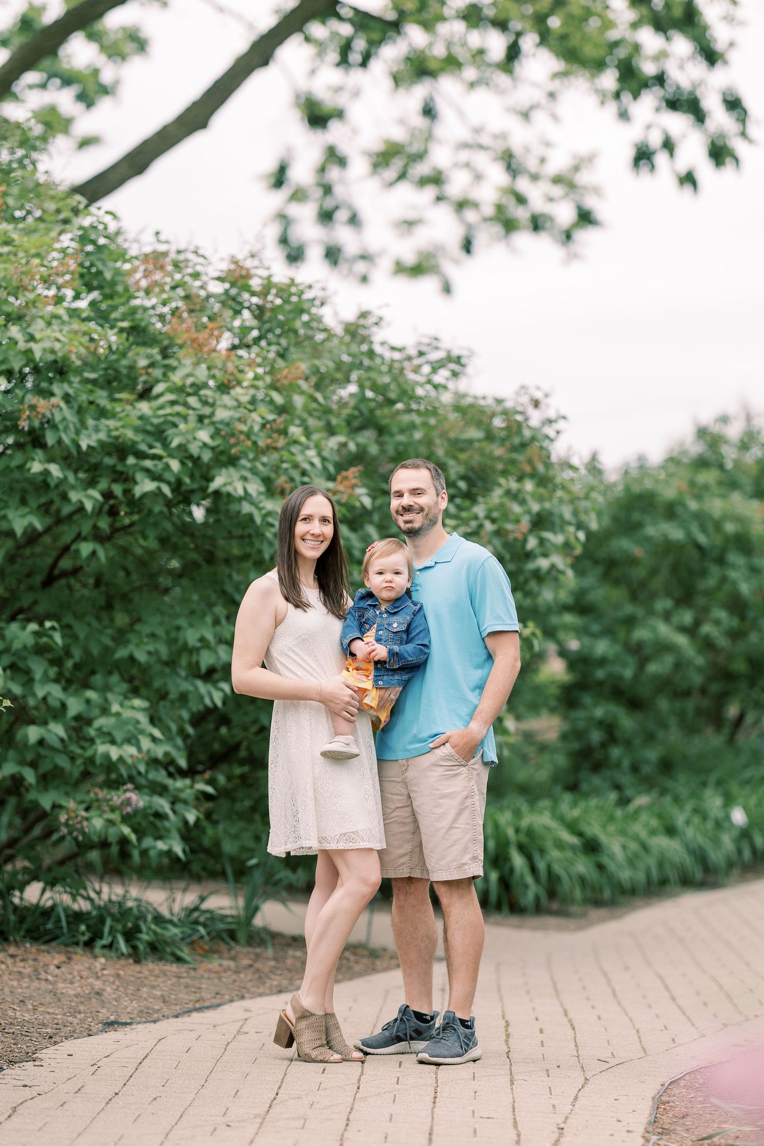 Smiling family of three posing on brick path surrounded by green summer foliage.