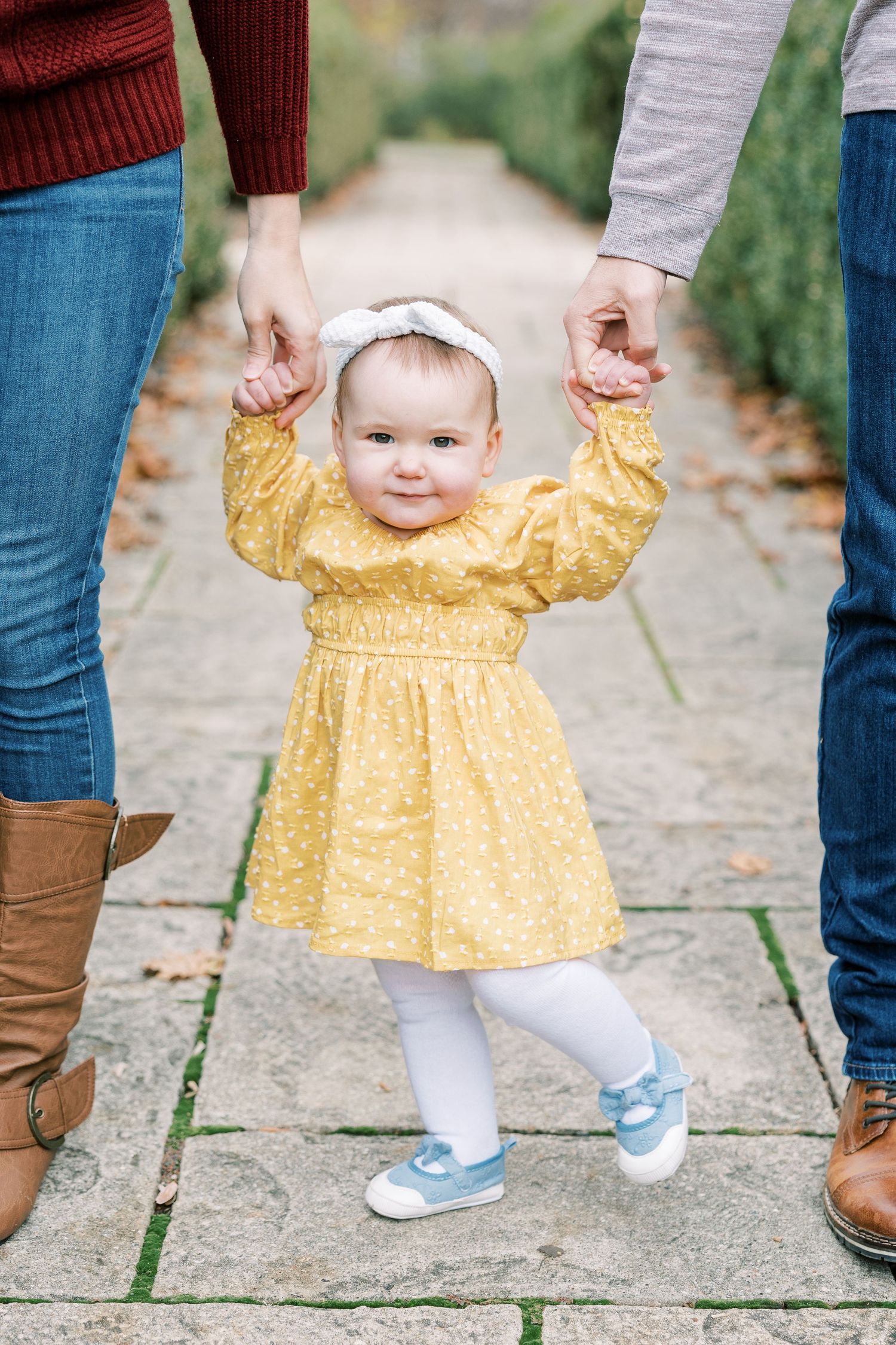 Baby girl in yellow dress holding parents hands while learning to walk on stone path.