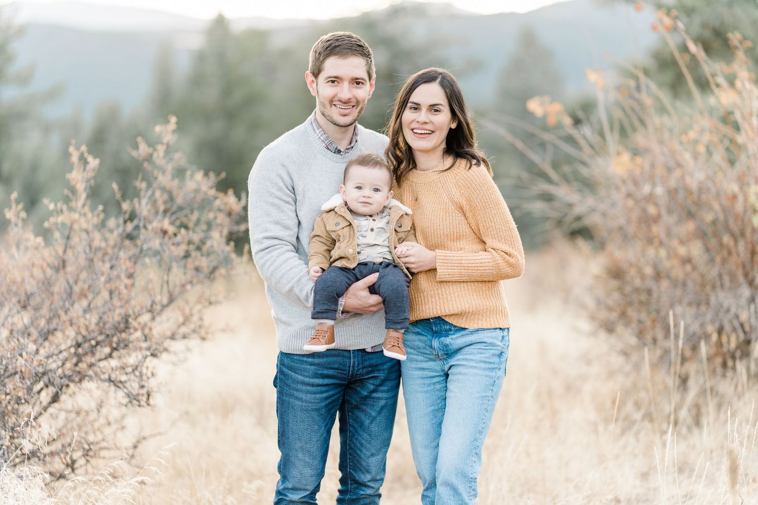 A young family of three poses together in a scenic field with mountains in the background.