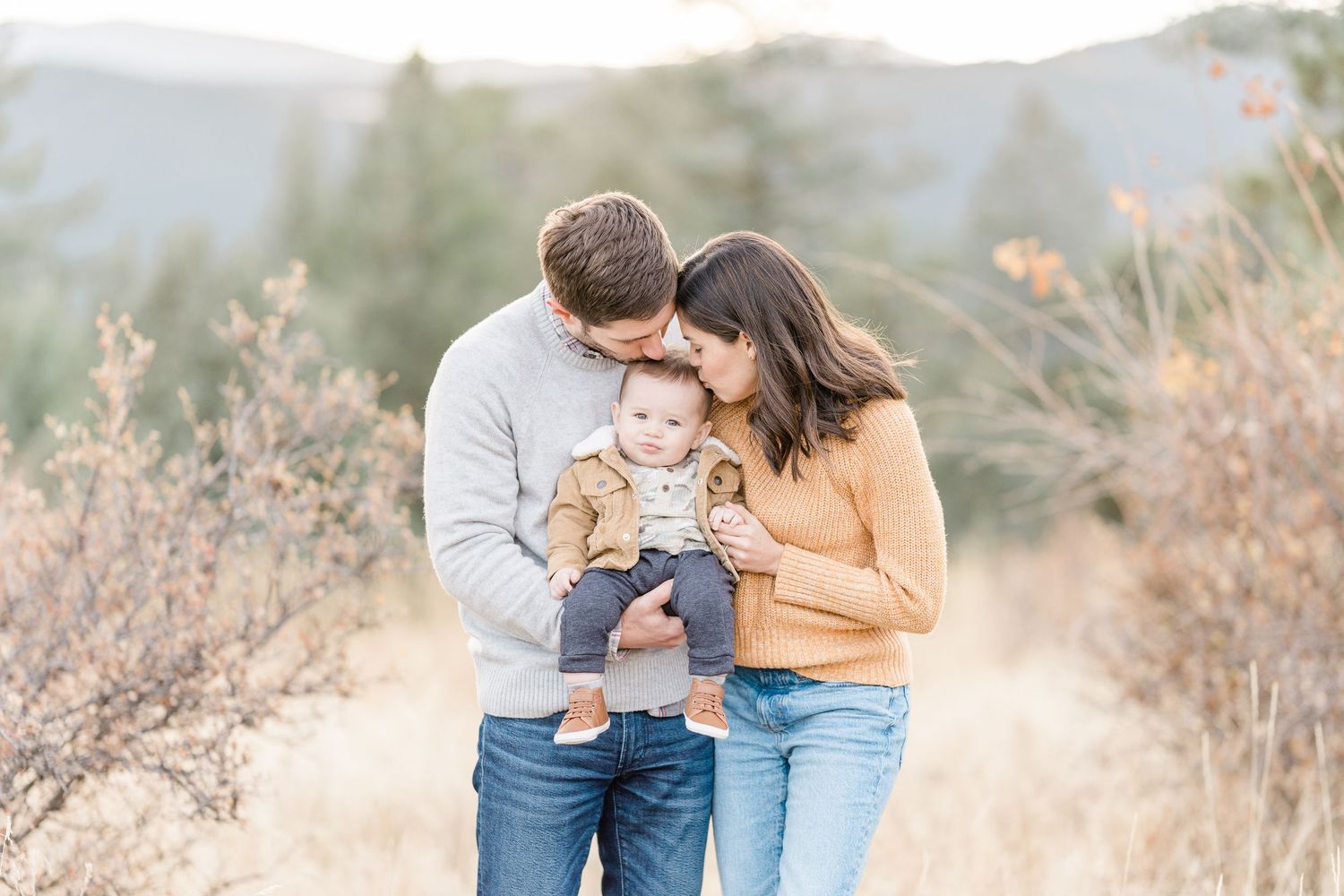 A couple kisses their baby outdoors in a golden field with mountain views during a fall family photo session.