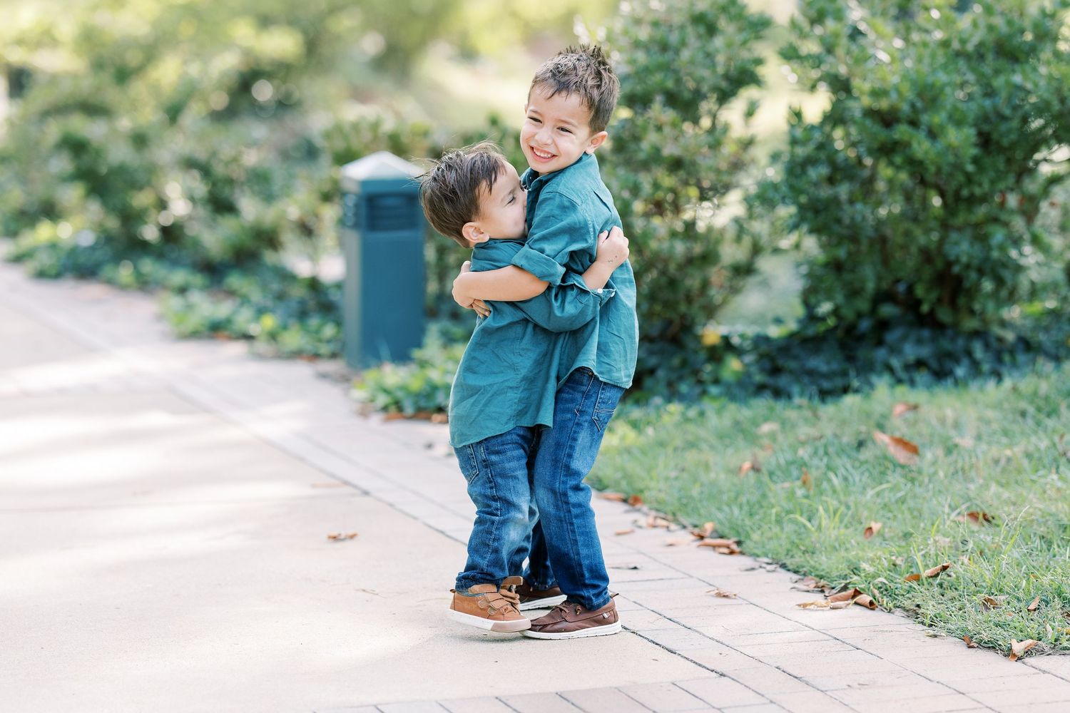 Two young brothers in matching teal shirts hugging each other on a sunny park pathway.