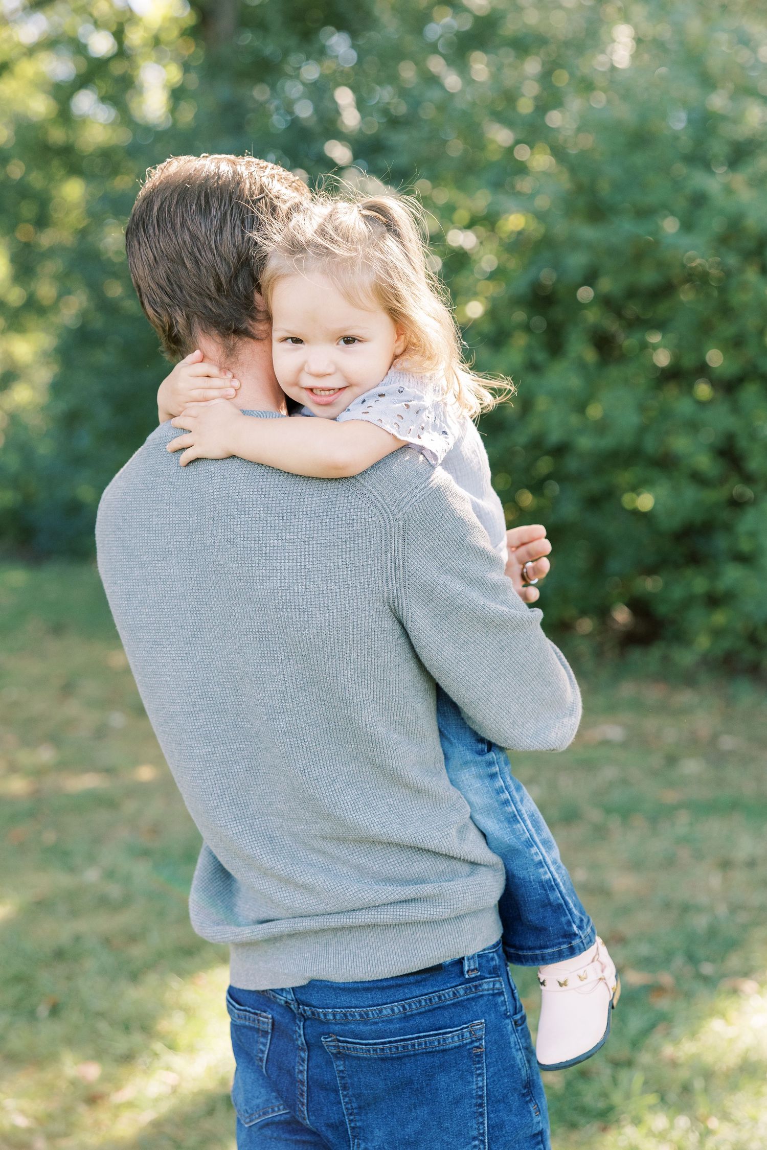 Smiling toddler girl hugs father from behind outdoors, wearing jeans and white shoes in sunny garden.