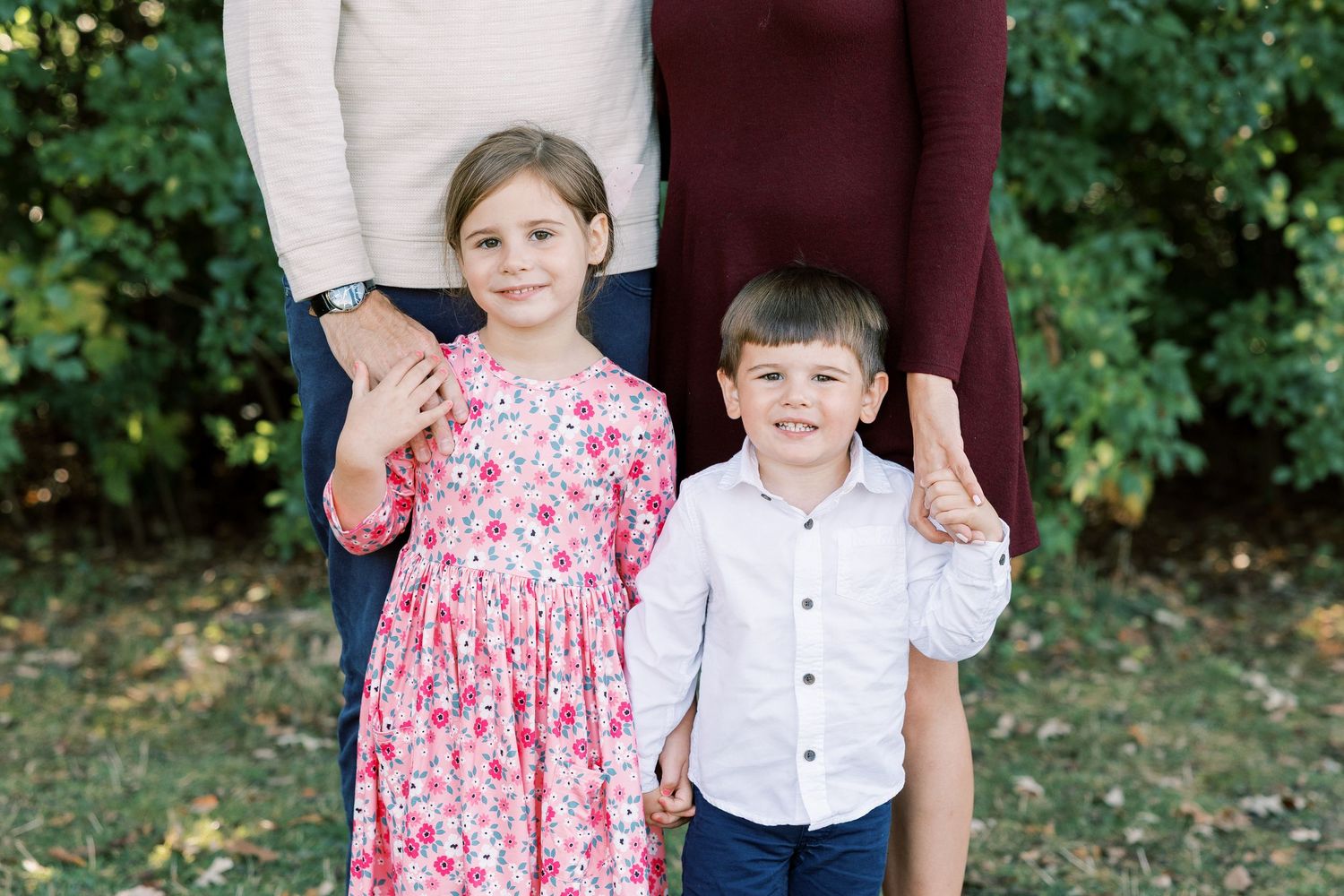 Two smiling children pose outdoors, girl in floral dress and boy in white shirt, both making peace signs.