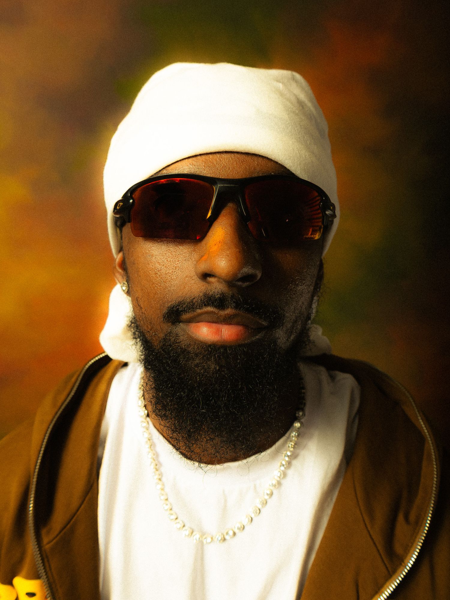 Man with white beanie, sunglasses, beard, and pearl necklace posing against colorful background.