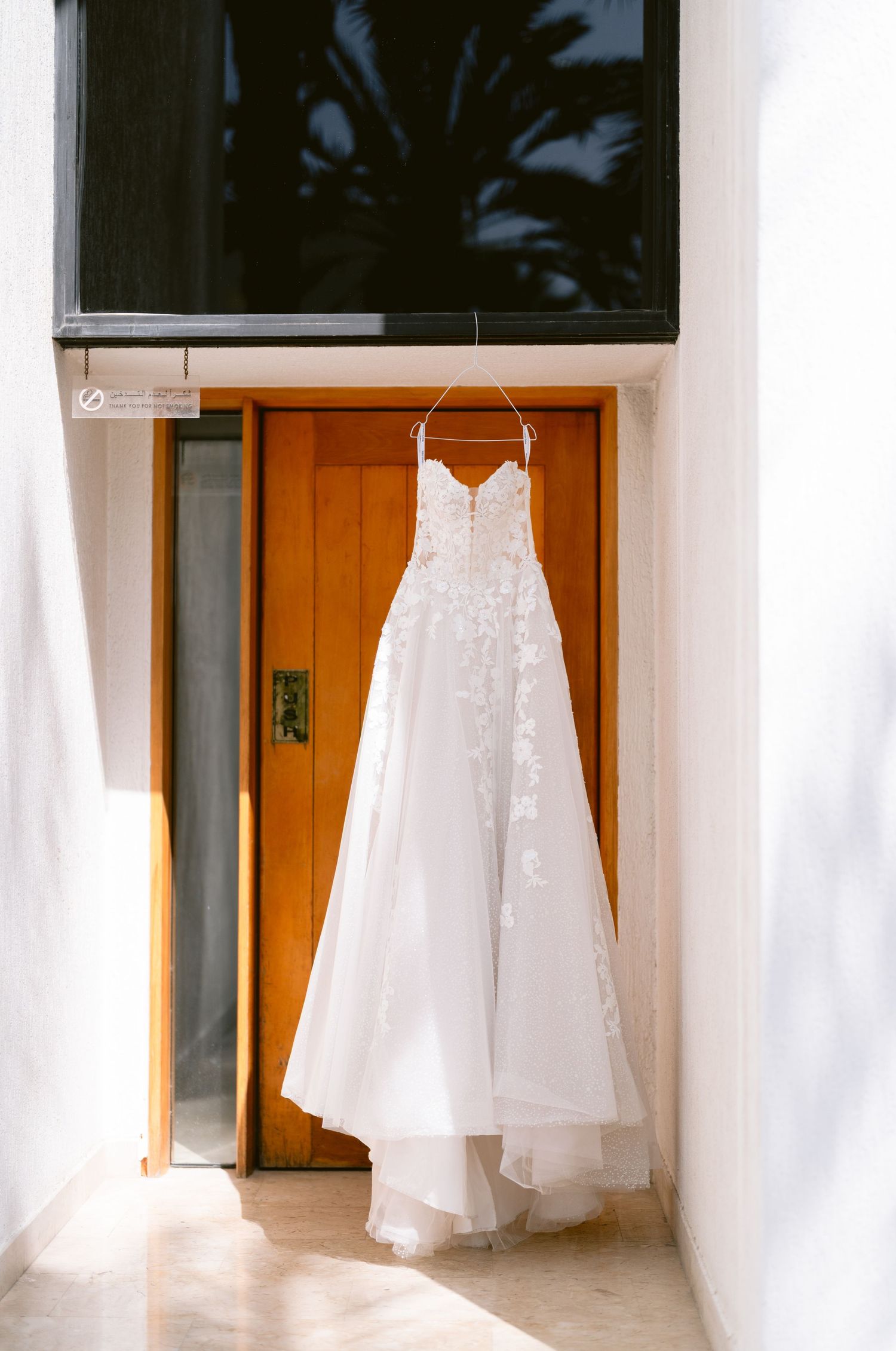 White strapless lace wedding dress hanging on a wooden door in a sunlit room.