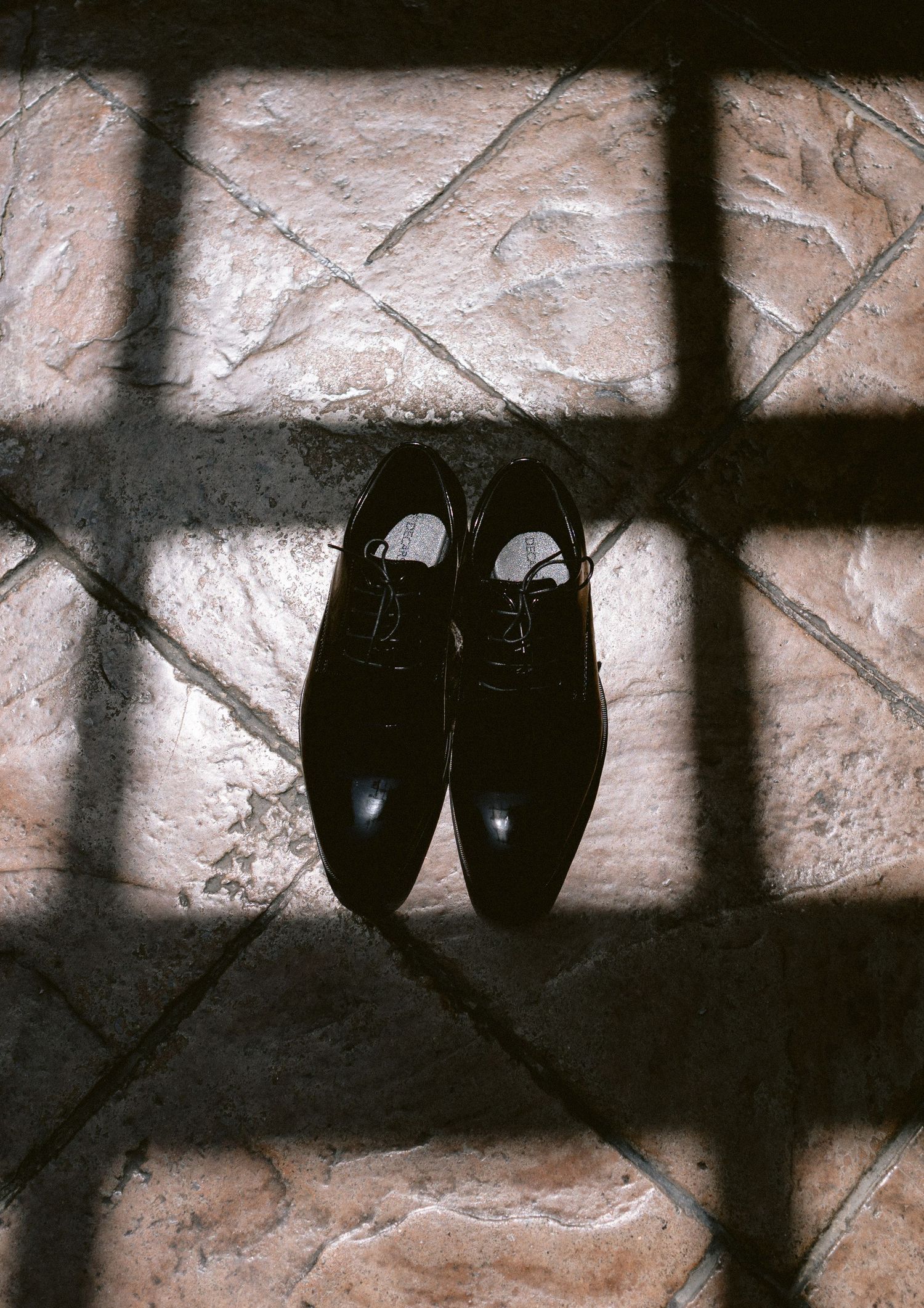 Shiny black dress shoes placed on rustic stone tile floor with dramatic shadow pattern from window light.