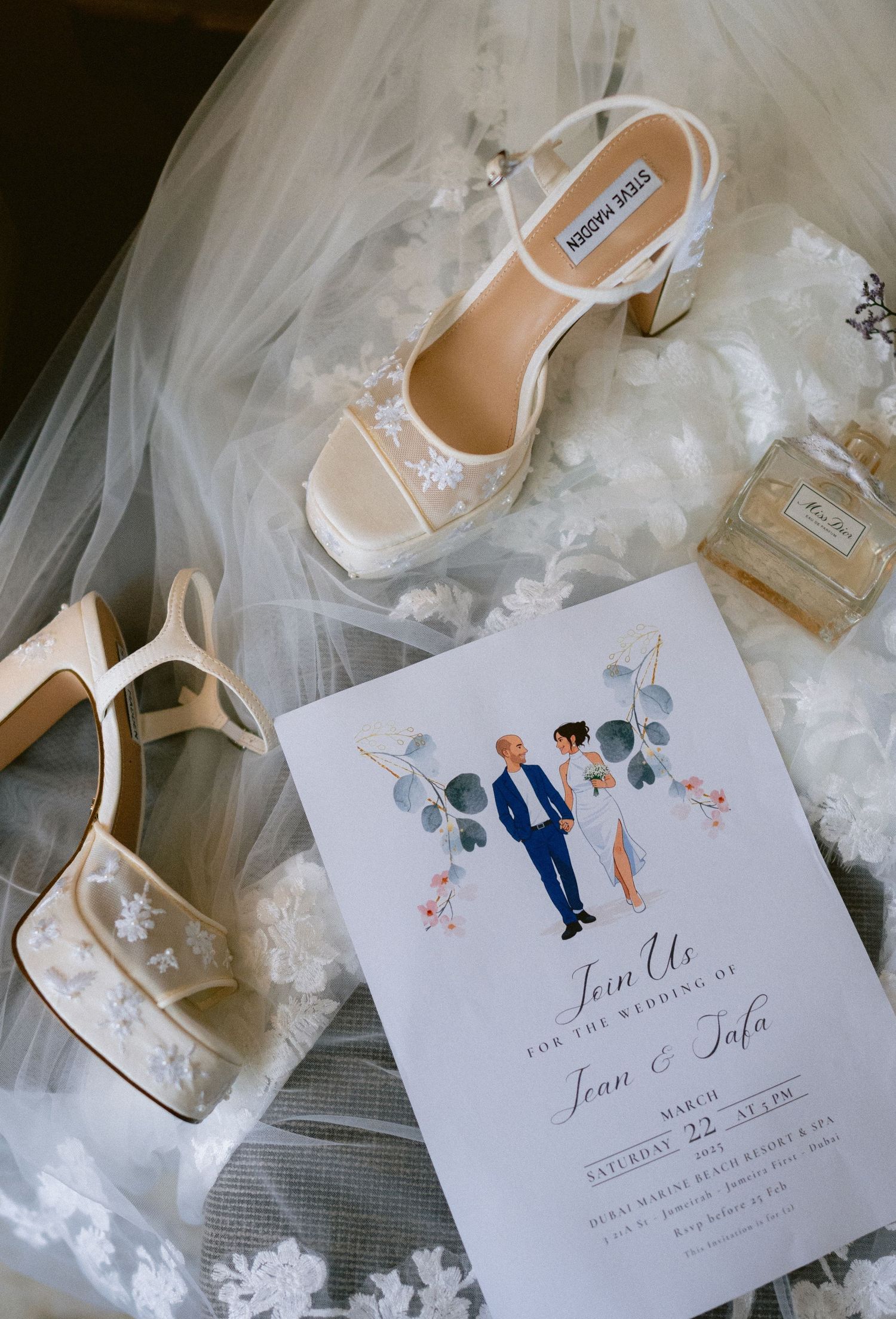 Elegant wedding flat lay featuring Steve Madden bridal heels, lace shoes, perfume, and a floral wedding invitation on tulle.