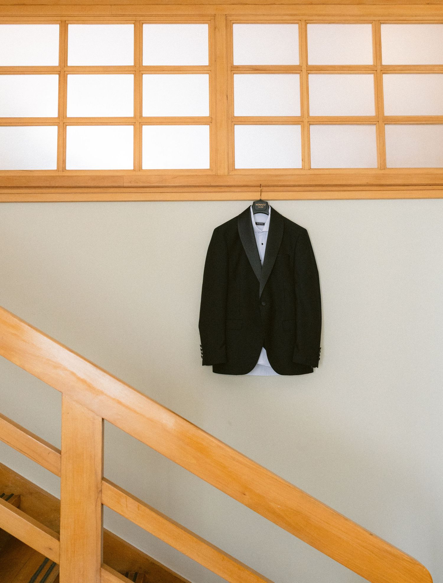 Black tuxedo jacket hanging on a hanger against a green wall near wooden staircase railing.