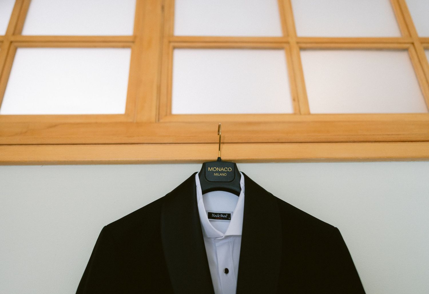 Black tuxedo jacket on hanger displayed against wooden framed window, Monaco brand label visible on formal suit.