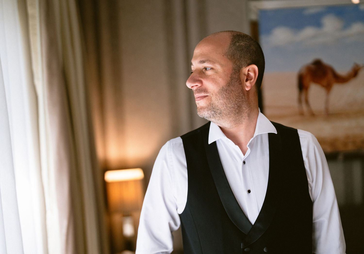 A bald man in a white shirt and black vest smiles while looking to the side in an elegant room.