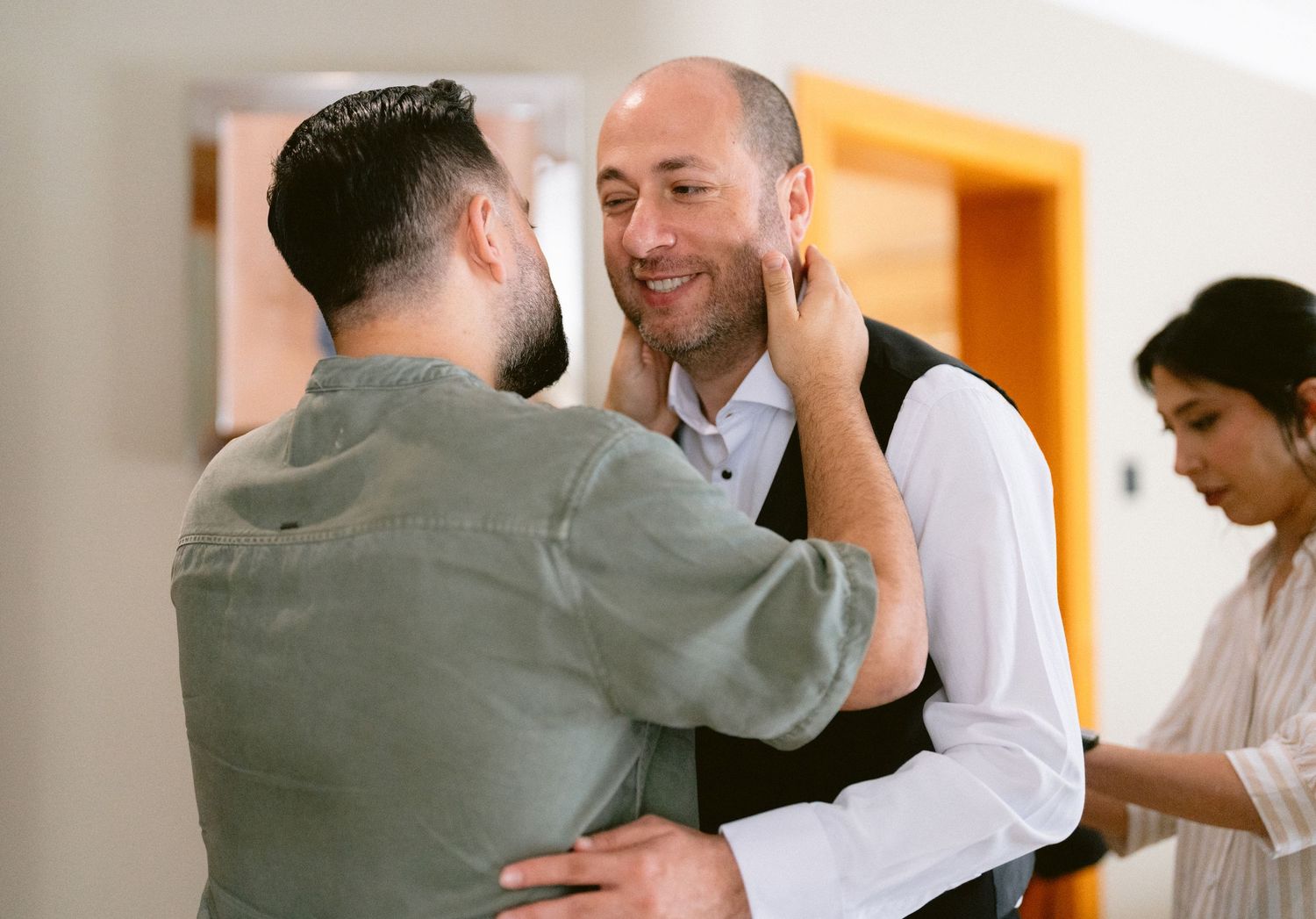 Two men share a warm embrace and smile at each other during a wedding celebration indoors.