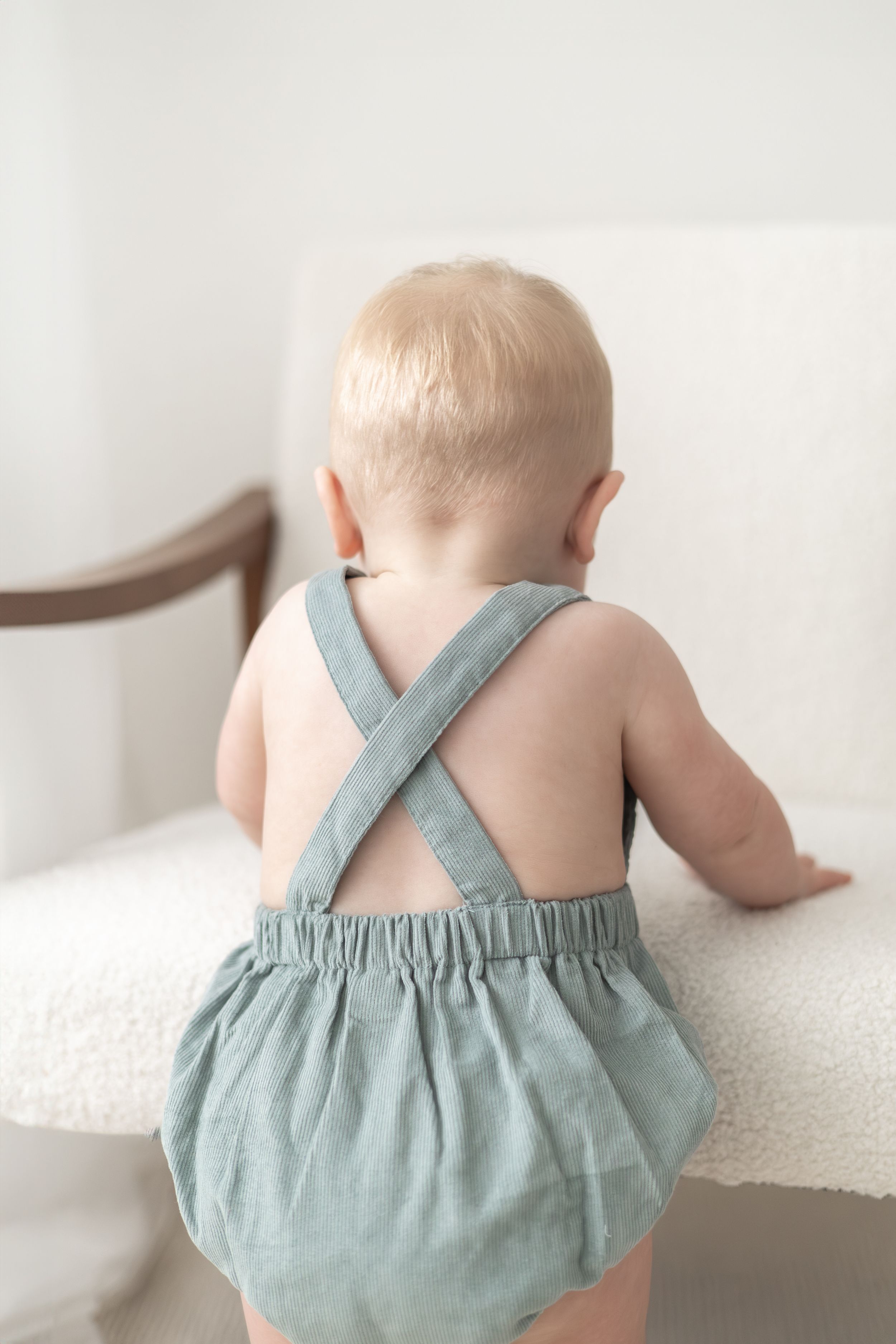 Baby in sage green linen romper with cross-back straps, standing at white chair. during birthday photoshoot Nottingham photography studio