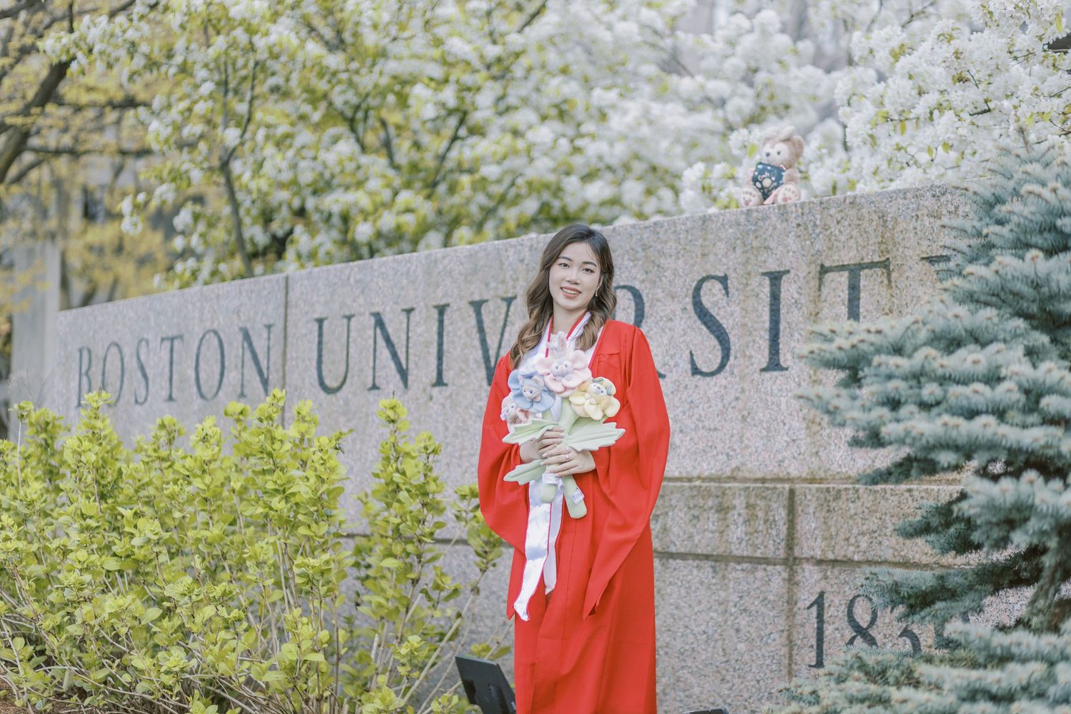 Boston University Graduate in red gown holding flowers poses in front of Boston University stone sign amid spring blossoms.
