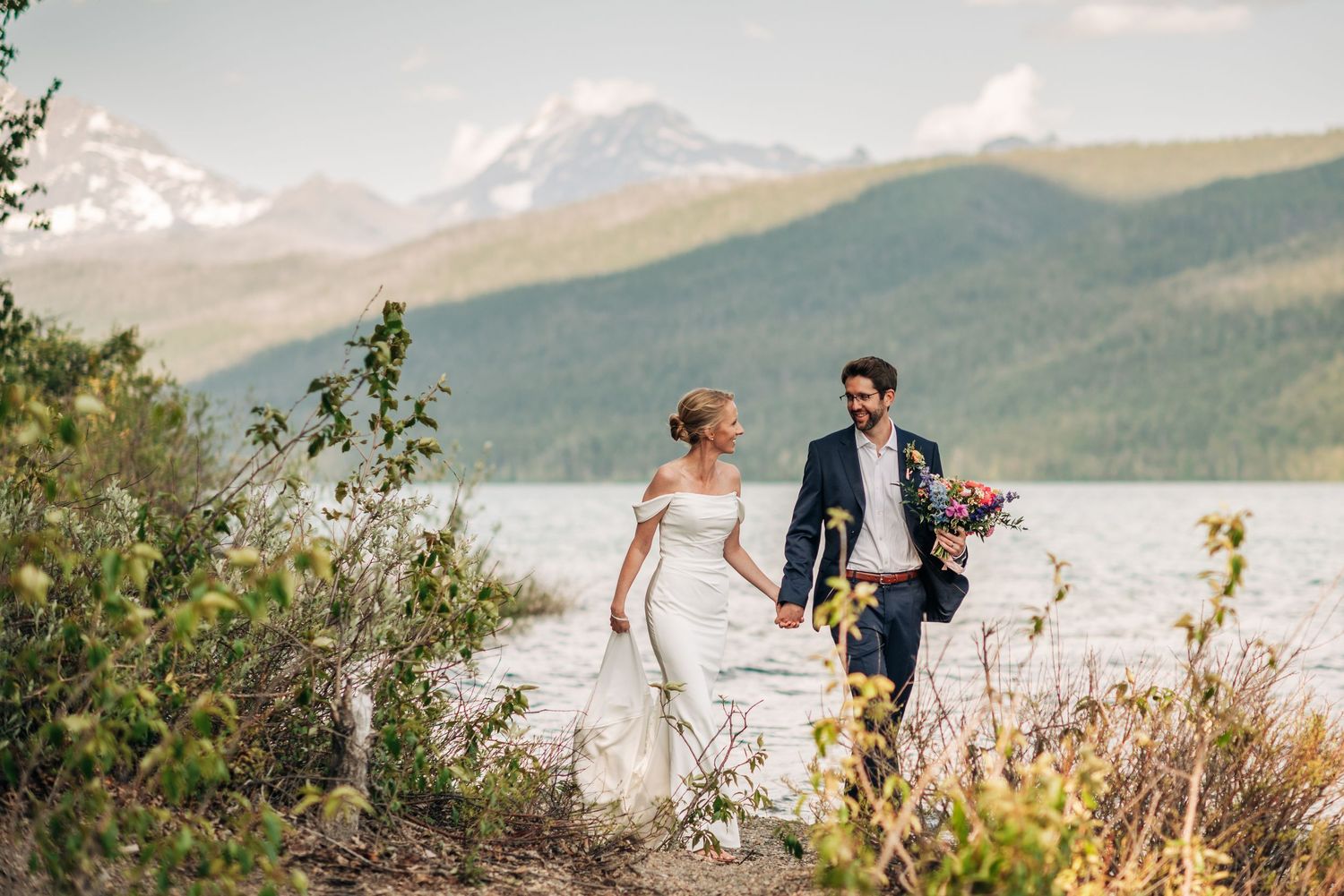 Bride and groom walk hand-in-hand along a scenic mountain lake shore with snow-capped peaks in the background.