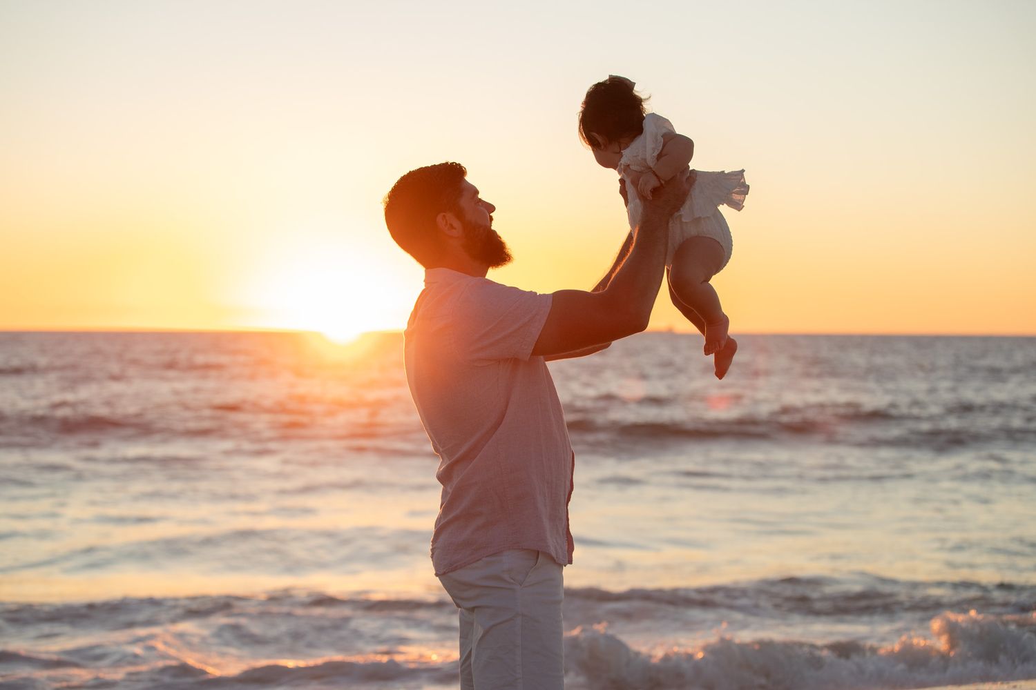 Father lifting baby joyfully at beach during golden sunset with ocean waves in background.