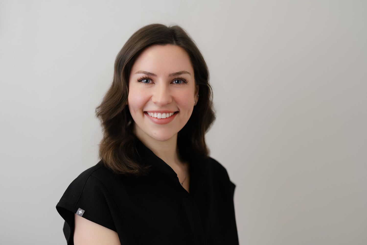 Professional headshot of a smiling young woman with brown hair wearing a black top against a light gray background.