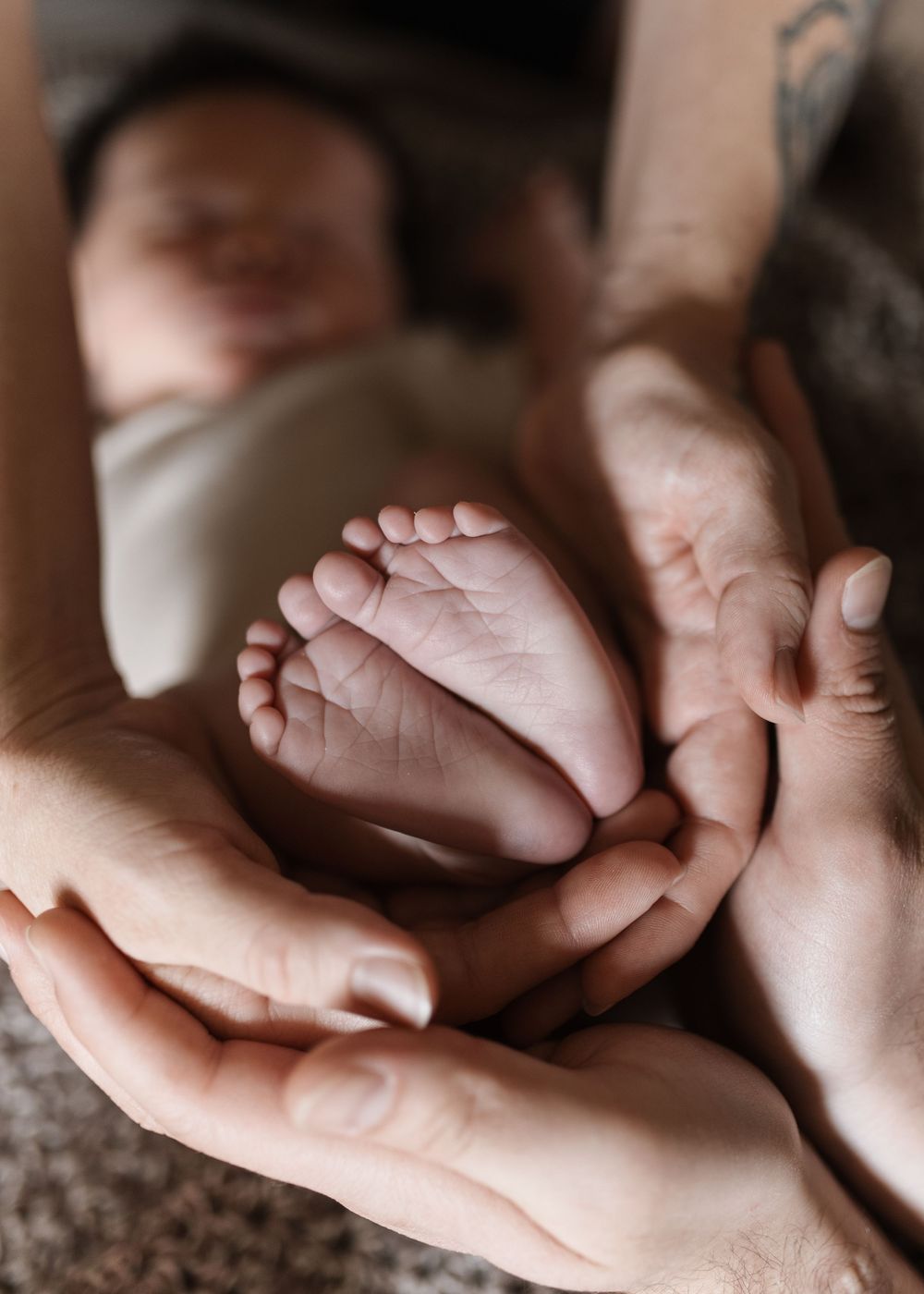 Parents cradling their newborn baby's tiny feet in their hands, with the sleeping infant visible in the background.
