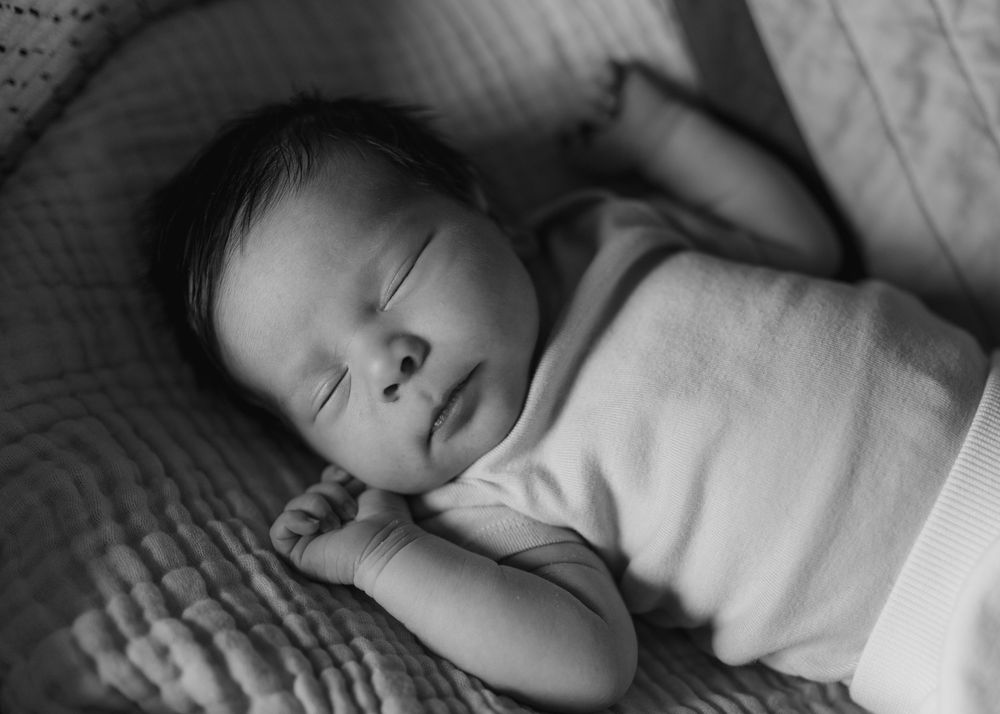 Black and white photo of a peaceful sleeping newborn baby swaddled in white blanket on a soft textured surface.