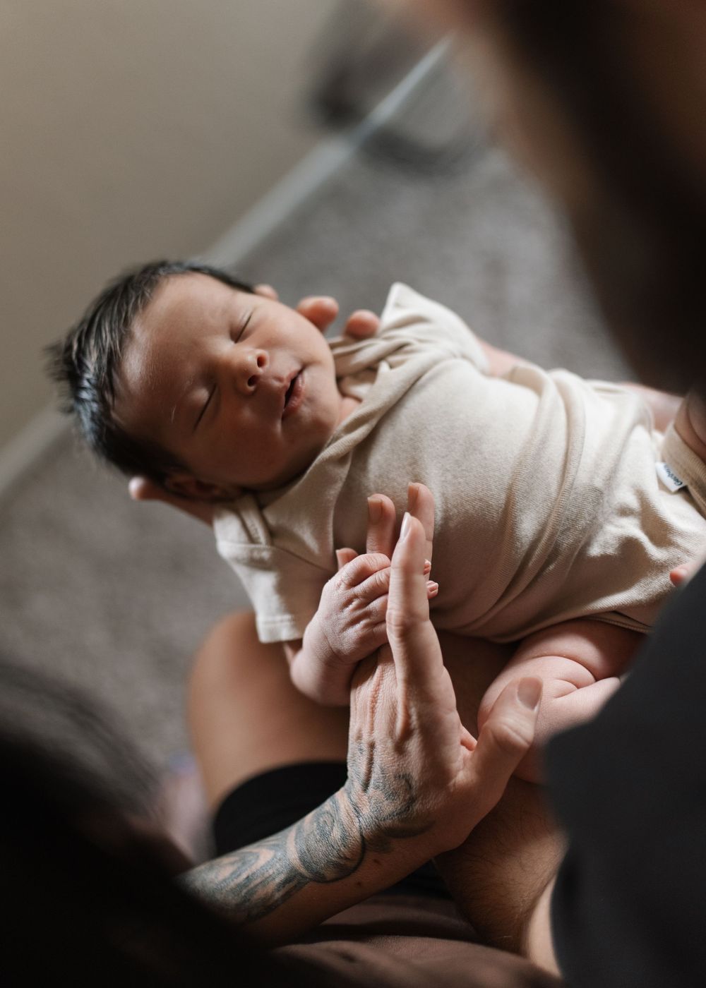 Newborn baby swaddled in cream wrap held gently by tattooed hands, eyes closed peacefully.