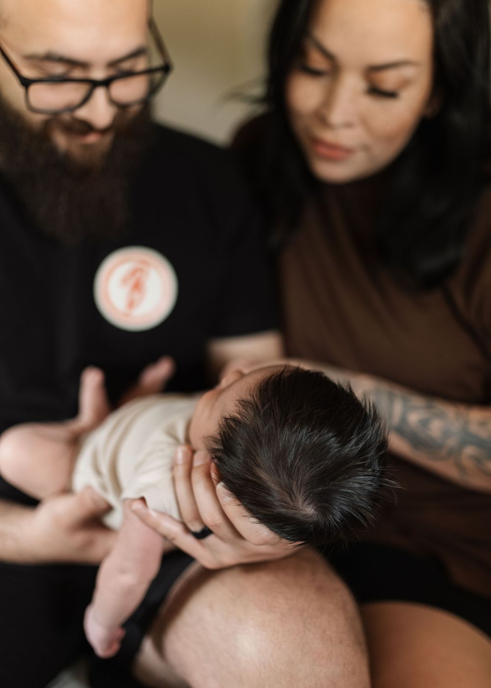 A bearded father with glasses and tattooed mother lovingly cradle their newborn baby together.