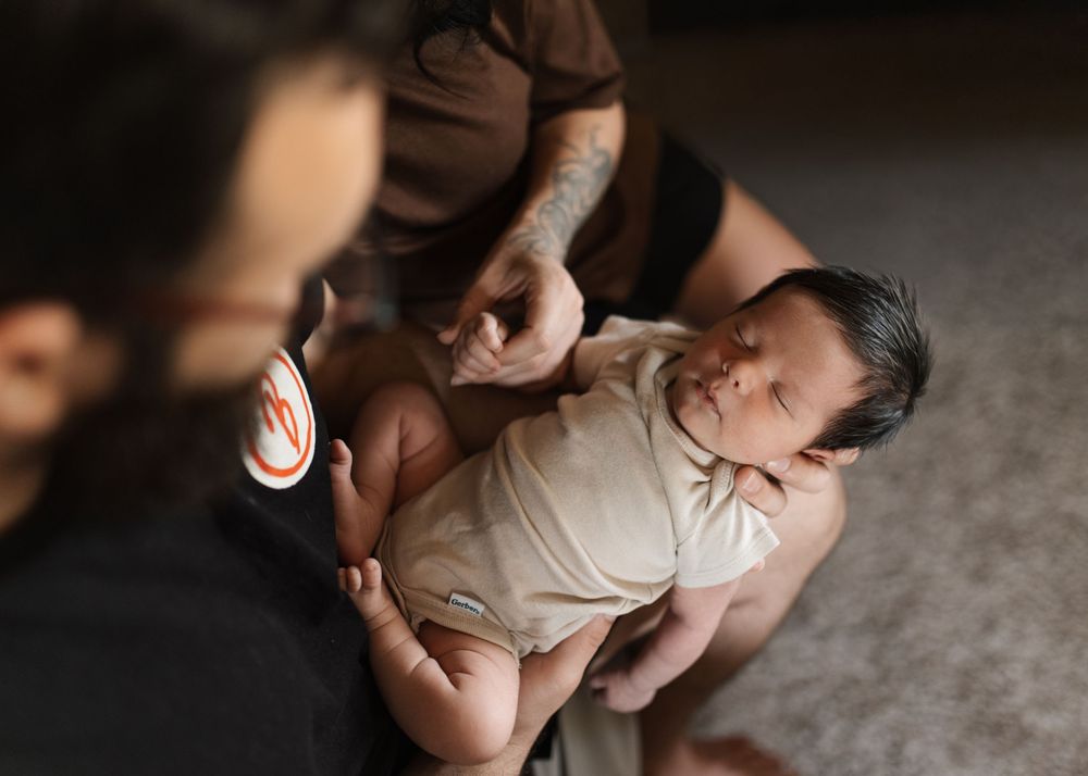 A tattooed parent gently holds a sleeping newborn baby dressed in a cream onesie during an intimate indoor moment.