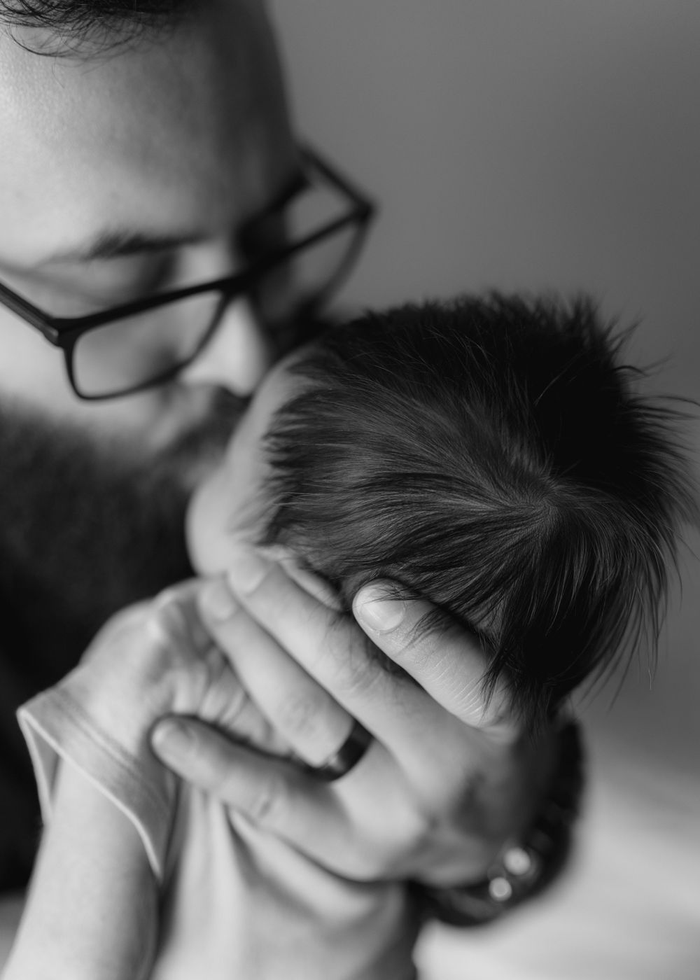 A bearded father with glasses tenderly kisses his newborn baby's head in black and white photo.