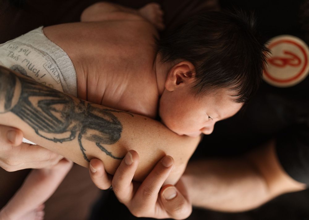 Tattooed adult hands gently holding a sleeping newborn baby close to their chest.