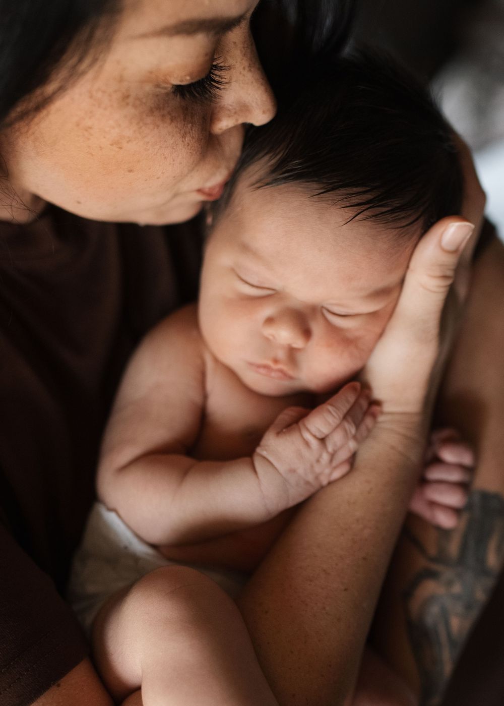 A mother tenderly holds her sleeping newborn baby close to her face in an intimate portrait.