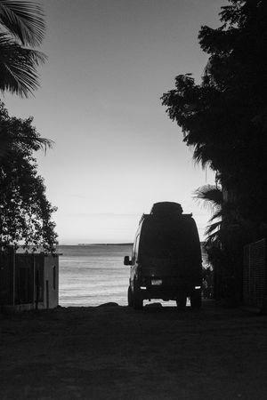 Black and white silhouette of a van parked between tropical trees with ocean view in background.