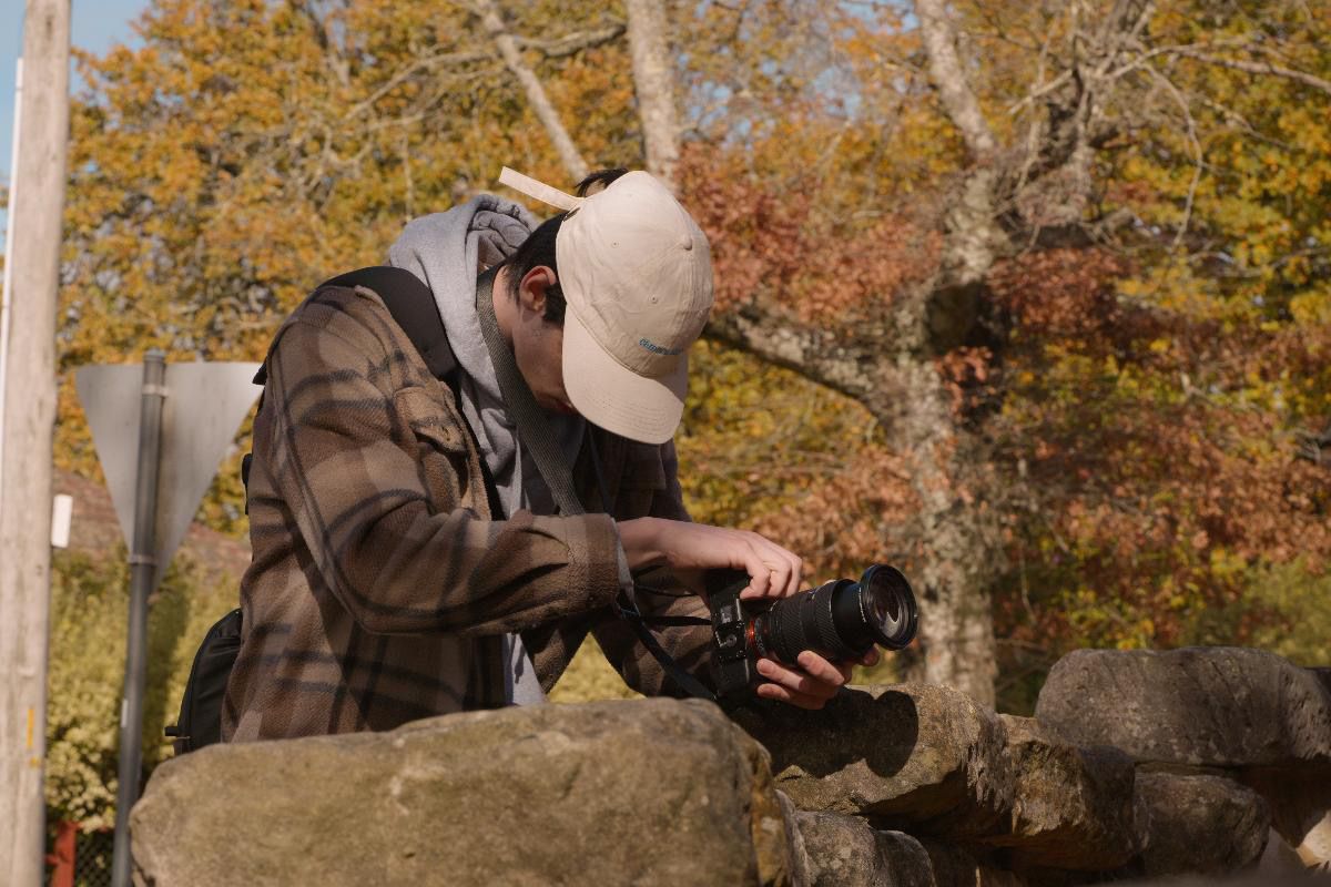 A photographer in a plaid shirt and cap reviews shots on a camera outdoors in autumn.