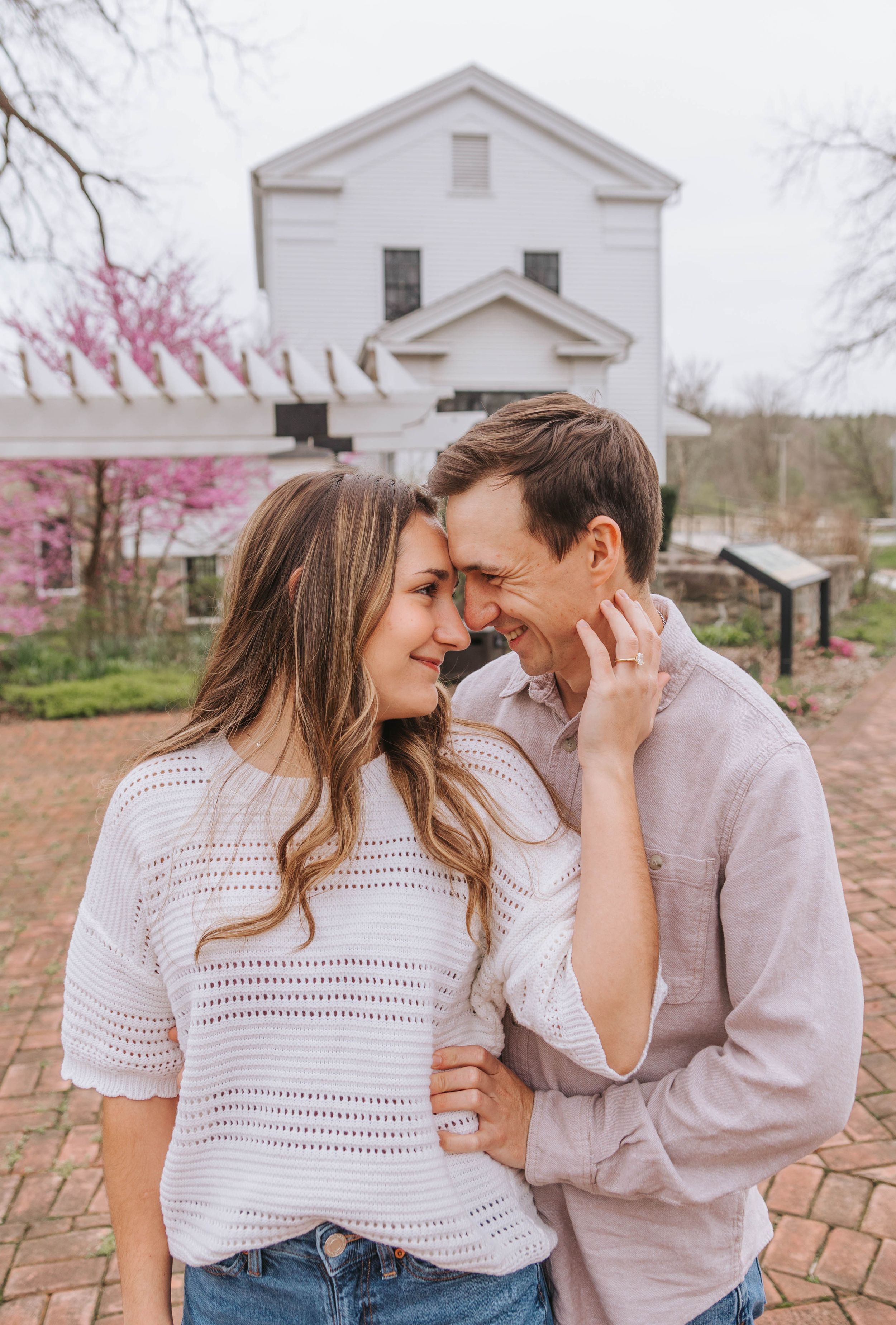 A smiling couple shares a tender moment outdoors near a white historic building with blooming redbud trees in spring.