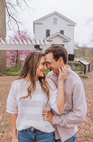 Happy couple leaning foreheads together smiling outdoors with white historic building and pink redbud tree behind them.