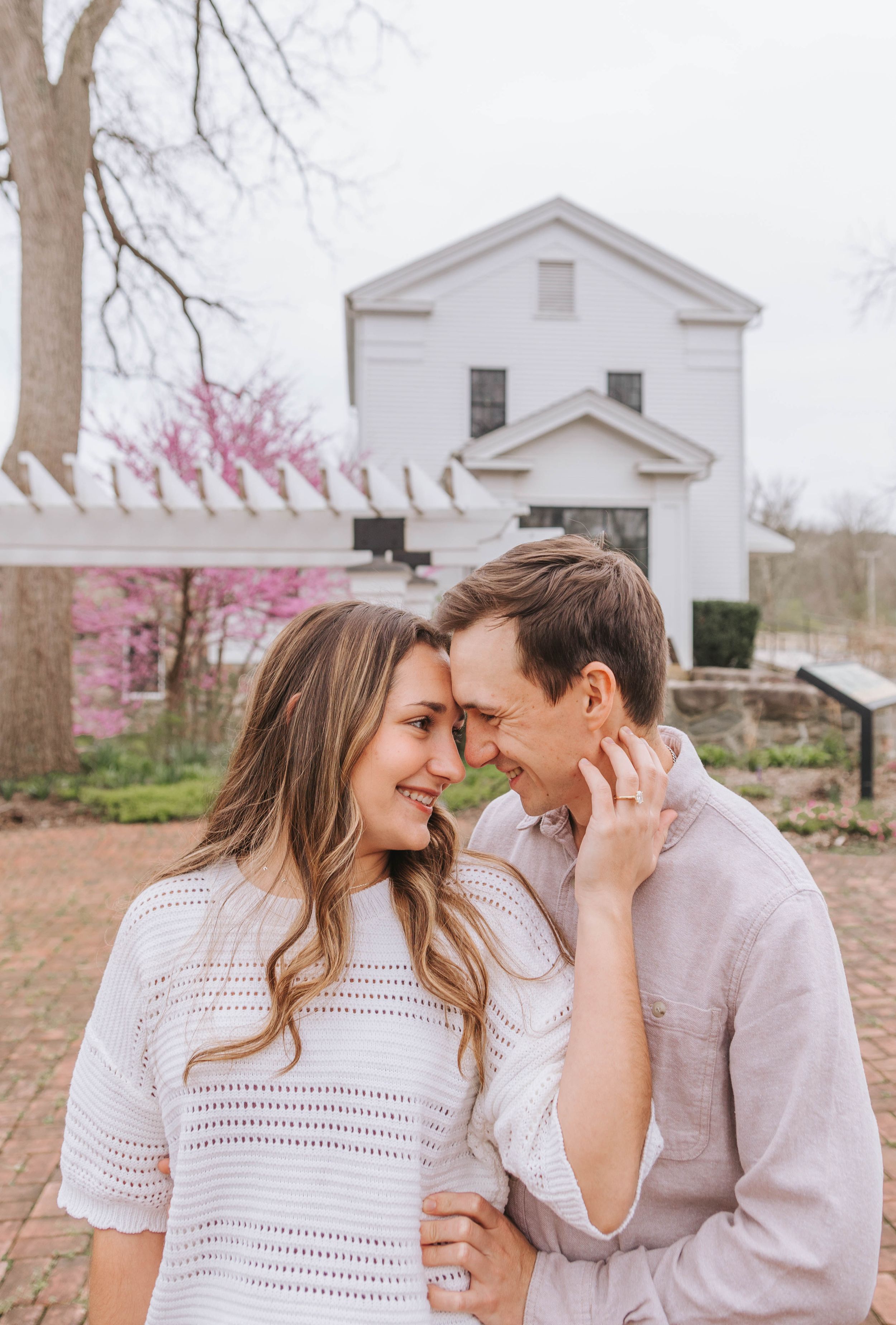 Smiling couple touching foreheads outdoors near white historic building with blooming redbud tree in spring.
