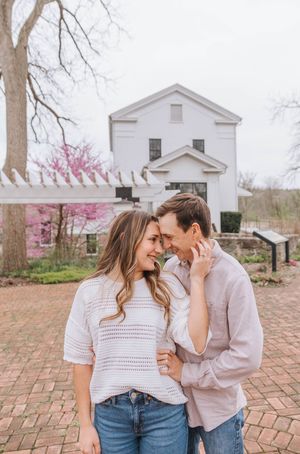 A couple shares a romantic moment outdoors near a white historic building with blooming redbud trees in spring.