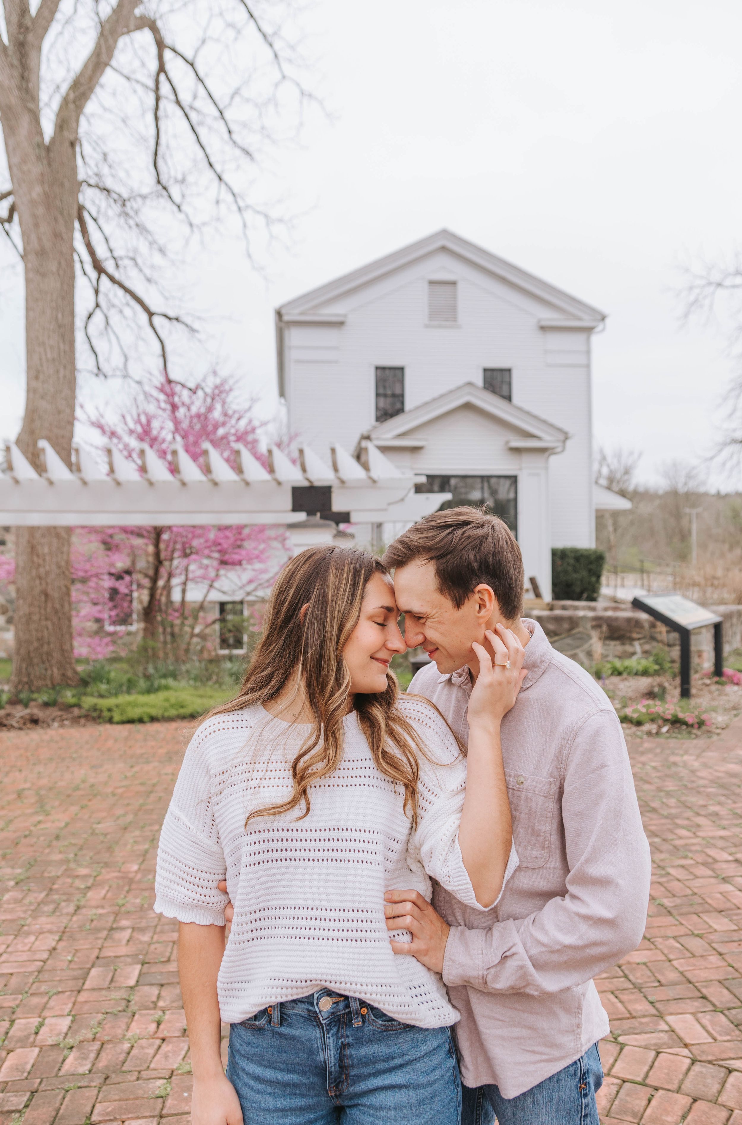 Couple sharing a romantic moment outdoors near a white historic building with pink blooming trees in spring.