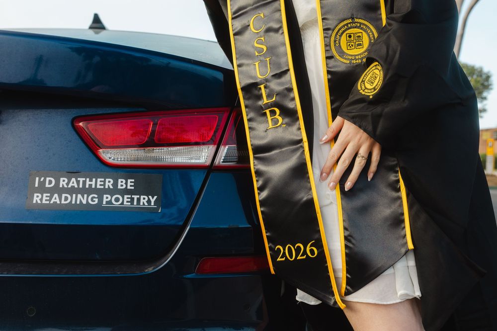 CSULB 2026 graduate in cap and gown beside a car with 'I'd Rather Be Reading Poetry' bumper sticker.