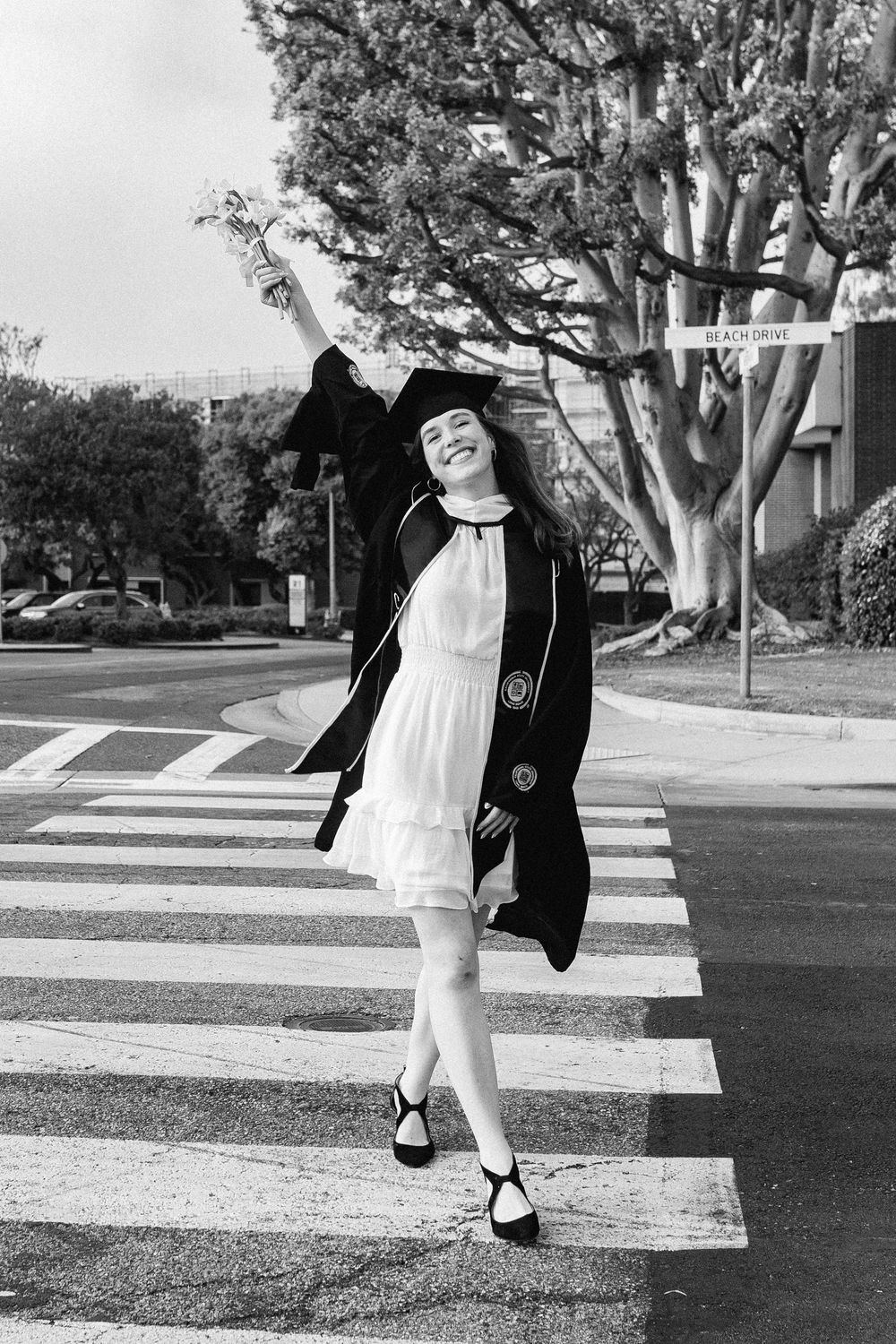 Graduate in cap and gown celebrates on a crosswalk in black and white photo.