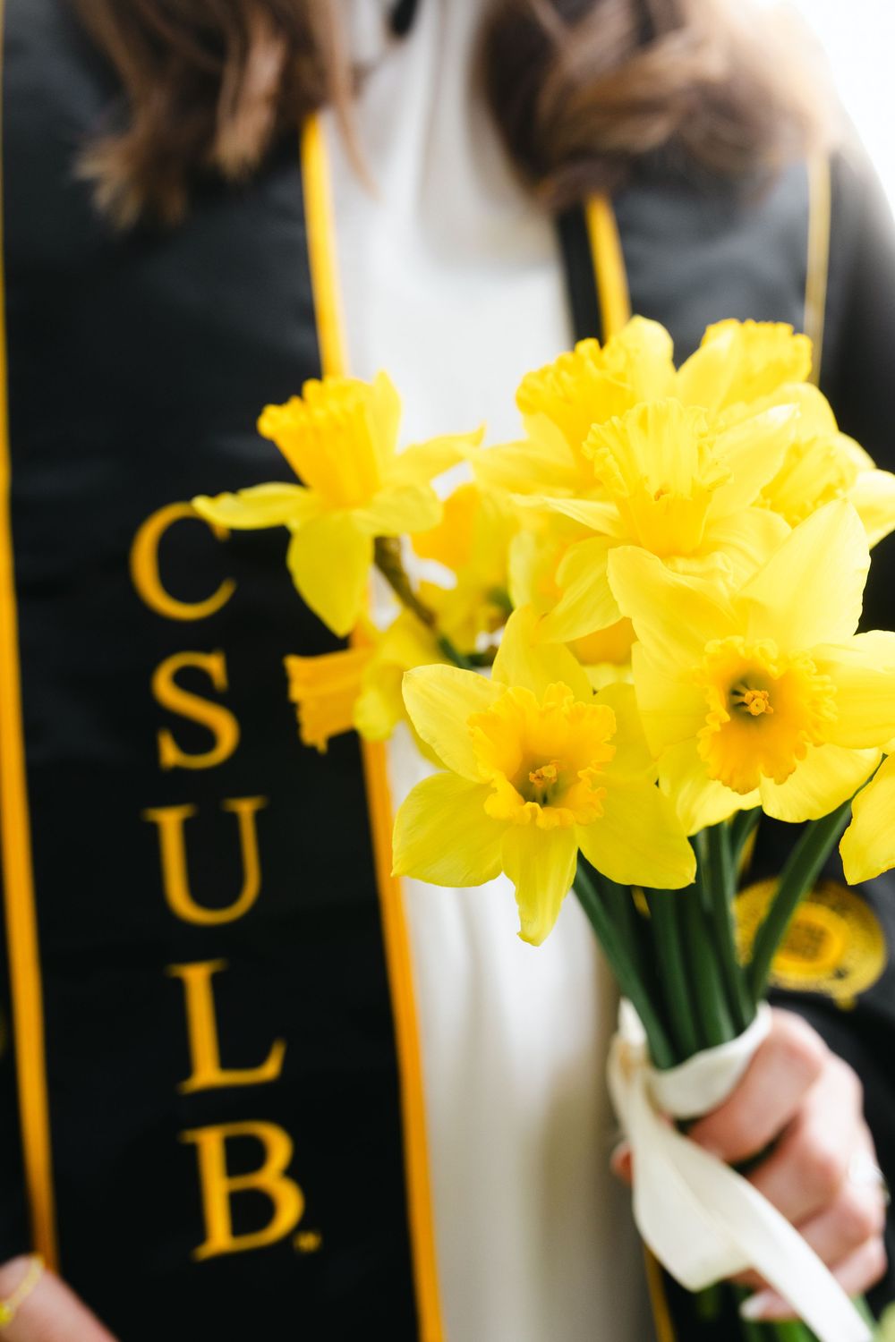 CSULB graduate holding bright yellow daffodils bouquet tied with white ribbon, wearing black and gold graduation stole.
